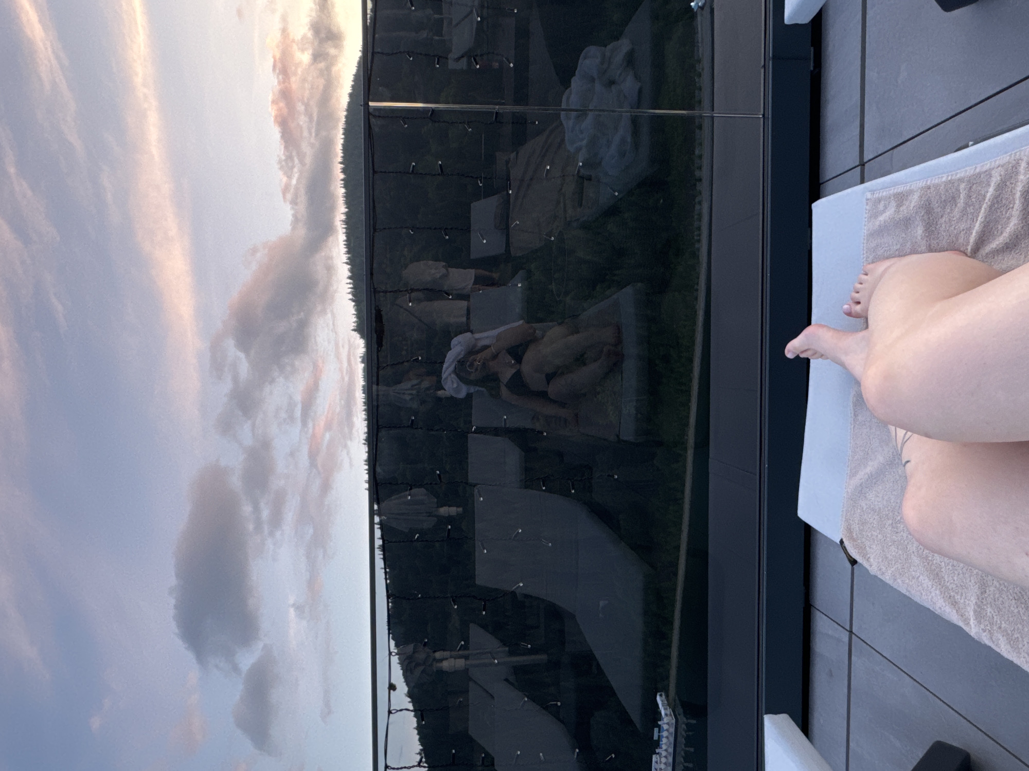 The image captures a serene sunset view from a rooftop lounger in Polianytsia, Ukraine. The foreground features the legs of a person resting on a beige towel atop a grey lounger. In the background, a reflective glass barrier separates the rooftop area from a lush, forested hillside. The glass reflects an array of lounge chairs, umbrellas, and a person dressed in a black bikini and white headscarf, also relaxing on a lounger. String lights are visible along the glass, adding a decorative element. The sky above is a soft blend of pink and blue hues, with scattered clouds and a distant treeline silhouetted against the twilight sky. The overall atmosphere is one of tranquility and relaxation, with the natural beauty of the landscape enhanced by the sunset's colors.