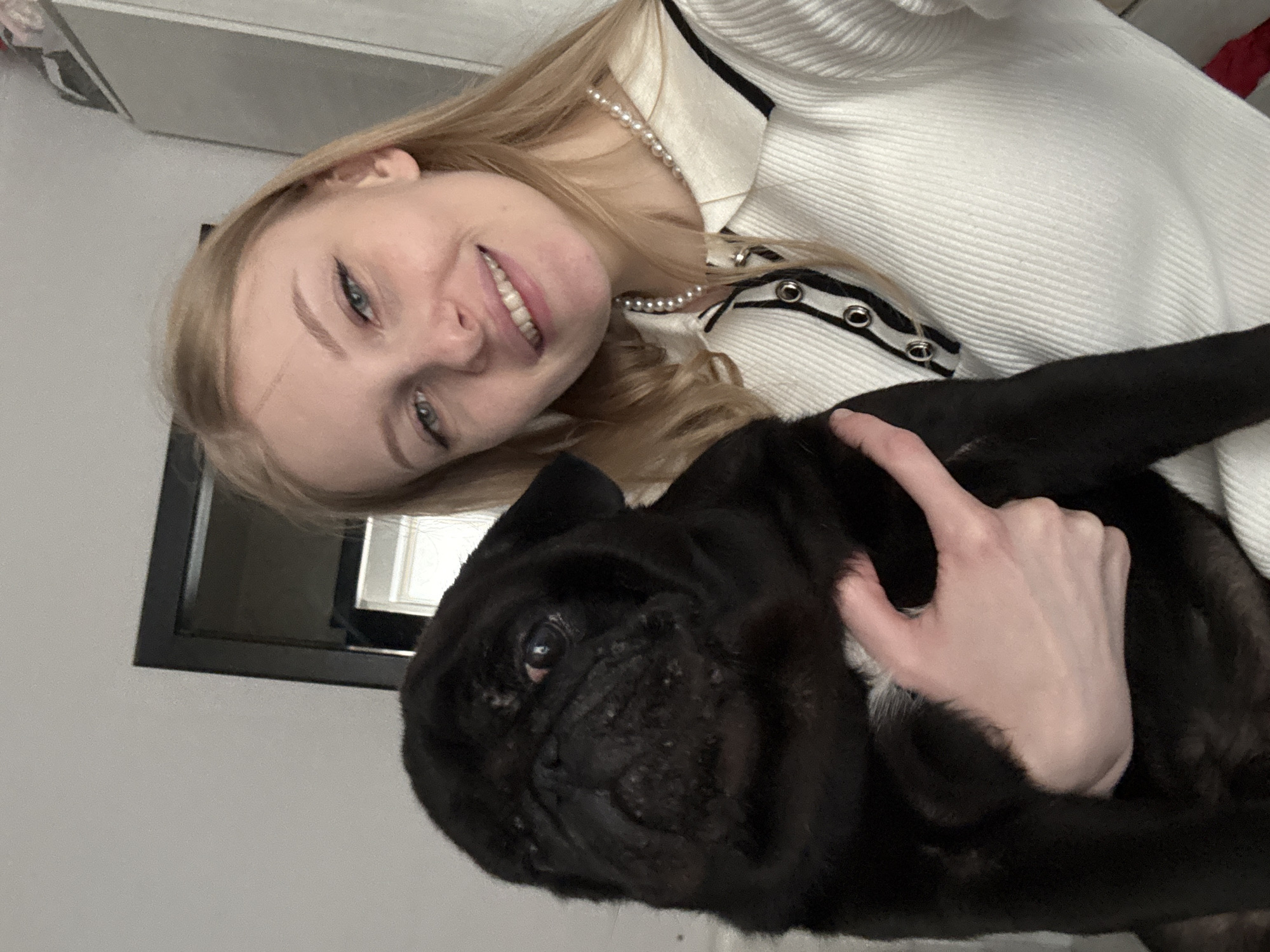 The image captures a young woman holding a black pug in an indoor setting, located in Perm, Russia.
The woman, with fair skin, light blonde hair parted on her right, and blue/green eyes, is smiling and looking directly at the camera. Her upper teeth show dental braces. She is wearing a white ribbed long-sleeved top with a white Peter Pan collar that has black trim and three decorative black buttons with silver rings down the placket. A single strand pearl necklace is visible around her neck. Her left hand is visible, holding the black pug.
The pug has a short, black coat with characteristic facial wrinkles. A small patch of white fur is visible on its chest. Its right eye appears slightly bloodshot or discolored.
The background consists primarily of a plain light-colored wall. To the upper right, a dark-framed object, possibly a mirror or picture frame, reflects a window with white frames. Further to the right, a section of a light-colored wardrobe or cabinet is visible. The room is brightly and evenly lit.