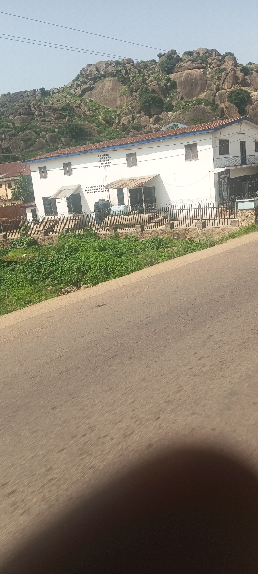 On Rukuba Road, Nigeria, a white building with a rusty metal roof stands before a rocky, vegetated hillside under a clear sky. The building has multiple windows and what appear to be air vents. A black metal fence with barbed wire lines the front of the property, which is elevated above a densely green, overgrown area. A paved road occupies the foreground, with a dark shadow obscuring the lower right corner of the image.