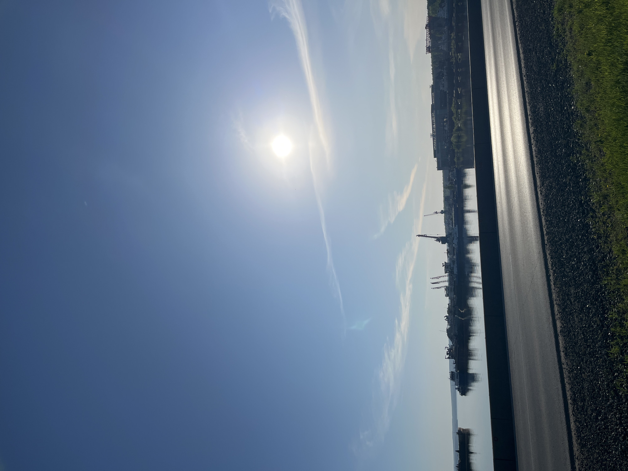 The image shows a view in Chaikovsky, Russia on a sunny day. The sky is predominantly blue with some thin clouds and the sun shining brightly. A paved road with a narrow strip of gravel shoulder runs along the right side of the frame, with green grass at the very edge. Beyond the road, there is water, reflecting the sky and what appears to be a port or industrial area on the opposite shore. Cranes and ships are visible in the water, as well as buildings along the shoreline. The angle suggests the photo was taken at a low angle, likely from near ground level.