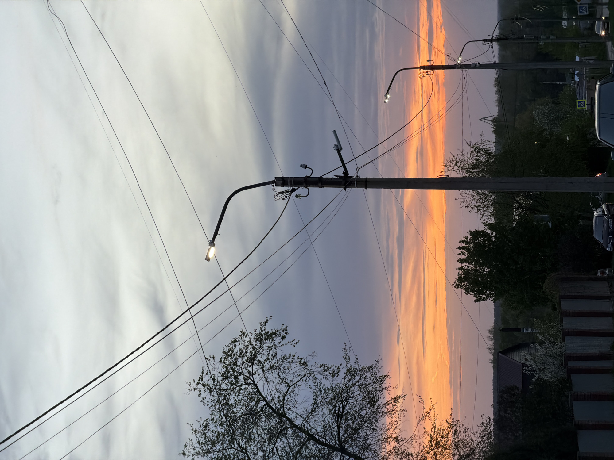 The image captures a street scene in Chaikovsky, Russia, at dusk or dawn. The sky is a mix of grey clouds and vibrant orange hues from the setting or rising sun. Overhead electrical wires crisscross the sky. Streetlights are visible, some illuminated. Trees with sparse foliage are in the foreground and midground. A house and a fence are in the lower right corner of the image. Cars and road signs are barely visible on the right.