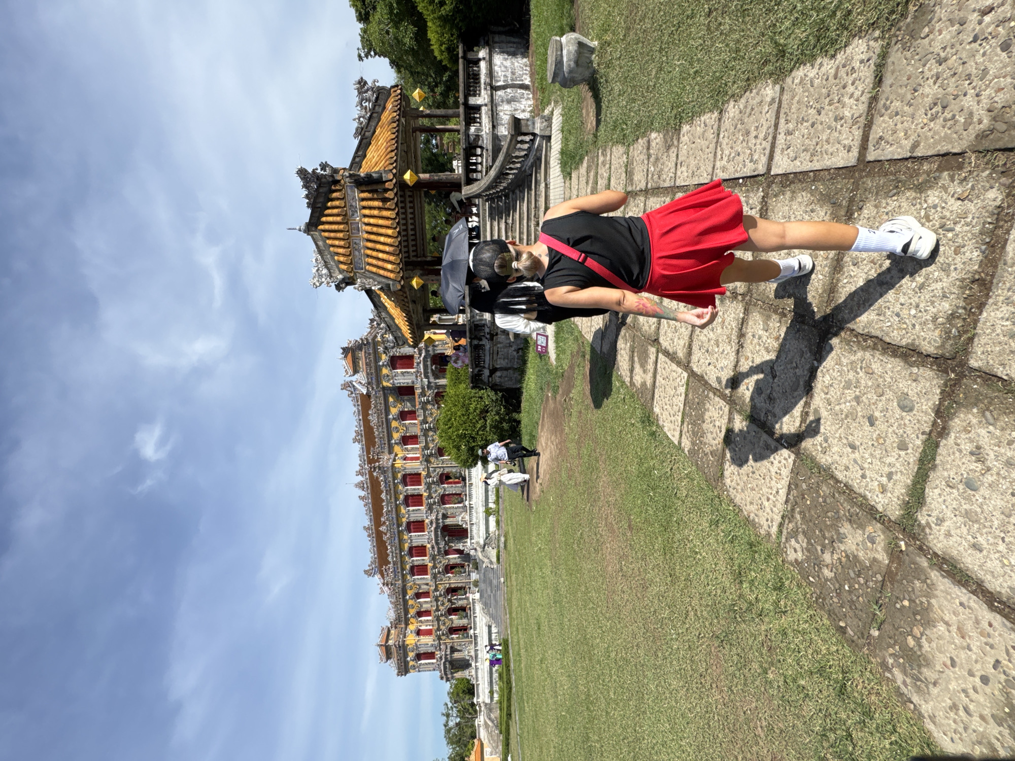 In Phú Xuân Ward, Vietnam, a woman with blonde hair pulled into a bun, wearing a black tank top, a red skirt, and white sneakers, walks on a stone pathway. She has a red strap across her shoulder and tattoos on her left arm. Her shadow is cast on the pathway. Behind her, another woman carries a black umbrella, and a third person is partially visible. In the distance, a large, ornate yellow and red building with red-trimmed windows stands under a partly cloudy blue sky. To the right of the pathway, a yellow-roofed pavilion with intricate carvings and yellow lanterns is situated on a raised platform with stairs. The surrounding area features a well-maintained green lawn and trees.