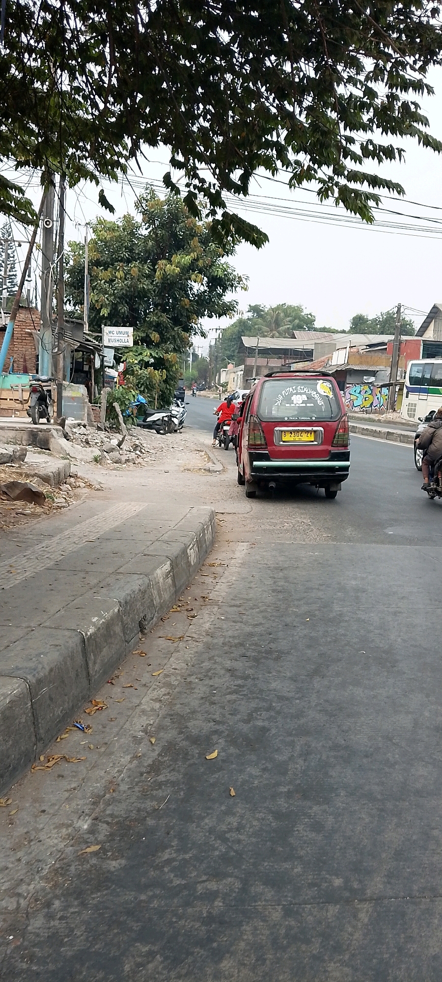 The image depicts a street scene in Bekasi, Indonesia, characterized by a road with a damaged sidewalk on the left and a lane of traffic on the right. A red car is parked or moving slowly on the right side of the road. Behind it, a motorcyclist in a red shirt is also visible. Further down the road, another motorcyclist is seen. The environment includes trees, overhead wires, and buildings along the side of the road, some of which appear to be shops or residences. There is debris and foliage scattered on the sidewalk and road. A sign that reads "VC UMUM MUSHOLLA" is attached to a pole on the left side of the image. The sky is overcast.