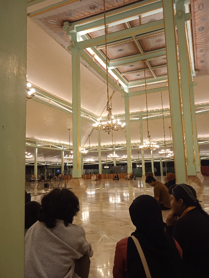 The image shows the interior of a large, open-plan building with a high ceiling and many ornate chandeliers hanging from it. The ceiling is decorated with light green and gold paint, and there are many large pillars supporting the structure. The floor is a light brown marble tile. A few people are present, sitting on the floor. The building is located at the coordinates -7.559, 110.783, which is the location of the Surakarta Palace in the city of Surakarta, Indonesia. The image captures a casual gathering of people enjoying the grandeur of the palace. The scene depicts a moment of peaceful contemplation within the palace's grand hall, where people are casually seated and engaged in conversation, seemingly enjoying the ambiance and the surroundings. It captures a glimpse into the daily life of the palace, showcasing a moment of peaceful interaction and enjoyment within its grand hall. The image is taken from a low angle, focusing on the people seated on the floor. The view is framed by one of the pillars, enhancing the perspective and emphasizing the height of the ceiling.