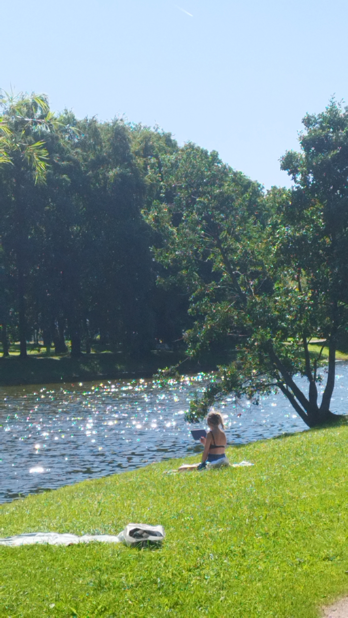 An outdoor, vertical, medium shot captures a person on a grassy embankment beside a body of water on a sunny day. The person, viewed from the back, sits with their legs crossed, wearing a black bikini. They are holding a tablet device. A white bag is visible in the foreground on the grass. The water is shimmering with sunlight. In the background, dense trees line the opposite bank of the water. The sky is clear blue with a faint contrail from an airplane. The overall impression is one of peaceful outdoor leisure. The location is likely a park or recreational area within Saint Petersburg, Russia, possibly along one of its many canals or the Neva River, given the typical urban park landscape of the city.