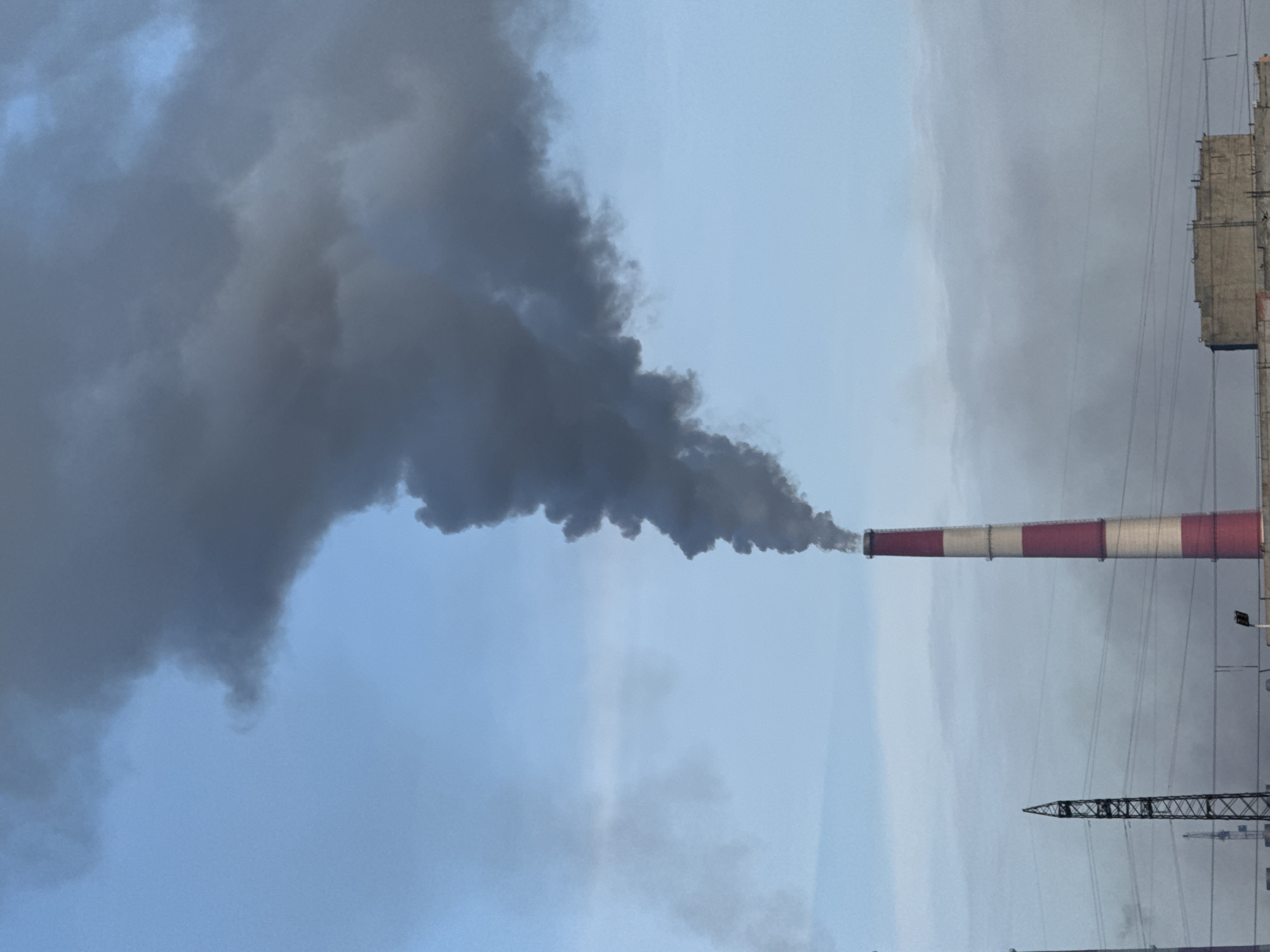 This image captures a panoramic view of an industrial area in Norilsk, Russia. The sky is a pale blue, partially obscured by wisps of gray smoke. Dominating the frame is a tall, red and white striped smokestack from which a thick plume of dark gray smoke billows upwards and spreads across the sky. To the left of the main smokestack, a tall, dark metal lattice tower stands against the sky, with power lines stretching horizontally across the scene. In the foreground, a low-lying concrete structure with visible rebar can be seen, suggesting ongoing construction or a derelict building. The overall impression is one of industrial activity set against a vast, and likely cold, northern sky.