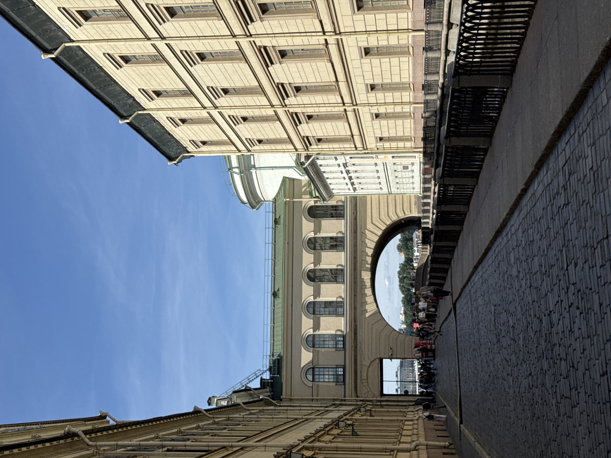 This is a street scene in Saint Petersburg, Russia, under a bright blue sky. On the right, a light-yellow building with a dark roof features a long facade with numerous windows. On the left, another light-yellow building with a detailed cornice runs along the street. The buildings are separated by a wide, arched passageway that bridges over a canal, visible in the distance. A cobblestone street occupies the foreground, leading towards the archway. Along the canal, a sidewalk with a decorative black metal fence runs parallel to the street. Several people are walking on the sidewalk and within the archway, some carrying bags. Trees line the far bank of the canal, and more buildings are visible in the background. The architecture suggests a historic district in the city center.