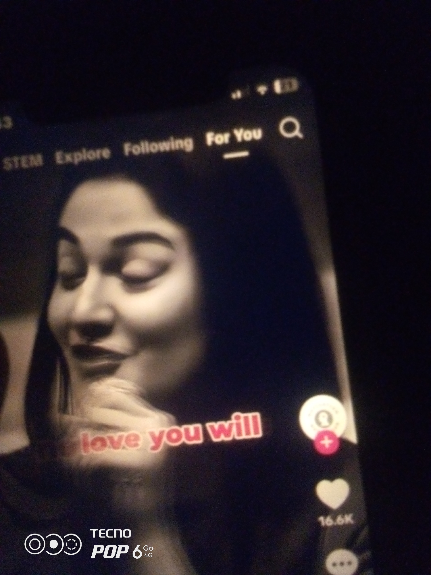 A blurred, low-light, close-up shot of a smartphone screen displays a social media video. The video features a young woman with her eyes closed and a subtle smile, her face partially illuminated. Text overlays on the video read "love you will". On the right side of the screen, icons for user interaction are visible: a profile icon with a plus sign, a heart icon with "16.6K" below it, and a three-dot icon. At the bottom left, the TECNO POP 6 4G logo is present. The background is dark and out of focus. There is no discernible information about the location in Howayi, Nigeria.