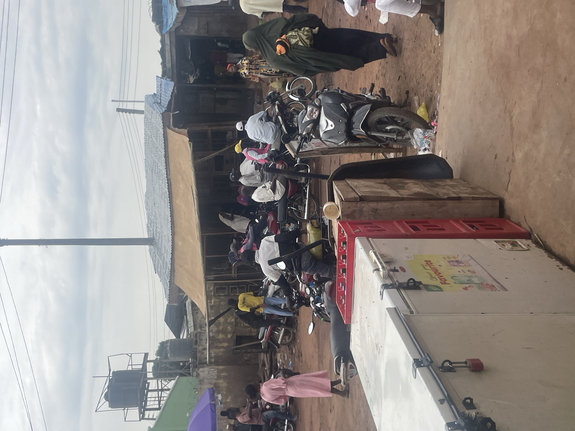 The image depicts a street scene in Lafia, Nigeria, likely a market or a busy thoroughfare, on an overcast day. In the foreground, a white chest freezer is visible, secured with a padlock, next to red crates. Beyond the freezer, a busy street unfolds with numerous motorcycles, some parked and others with riders. People are seen interacting and moving around. A woman in a pink dress is walking away from the viewer, and further back, a woman in a dark hijab with a baby strapped to her back is walking towards the right of the frame. Several men are present, some on motorcycles, and others standing or walking. Structures with corrugated metal roofs are visible in the background, characteristic of informal settlements or marketplaces. A water tank on a metal platform is also noticeable in the distance. The ground appears to be dirt or gravel. The overall impression is one of daily life and commerce in a Nigerian urban setting.