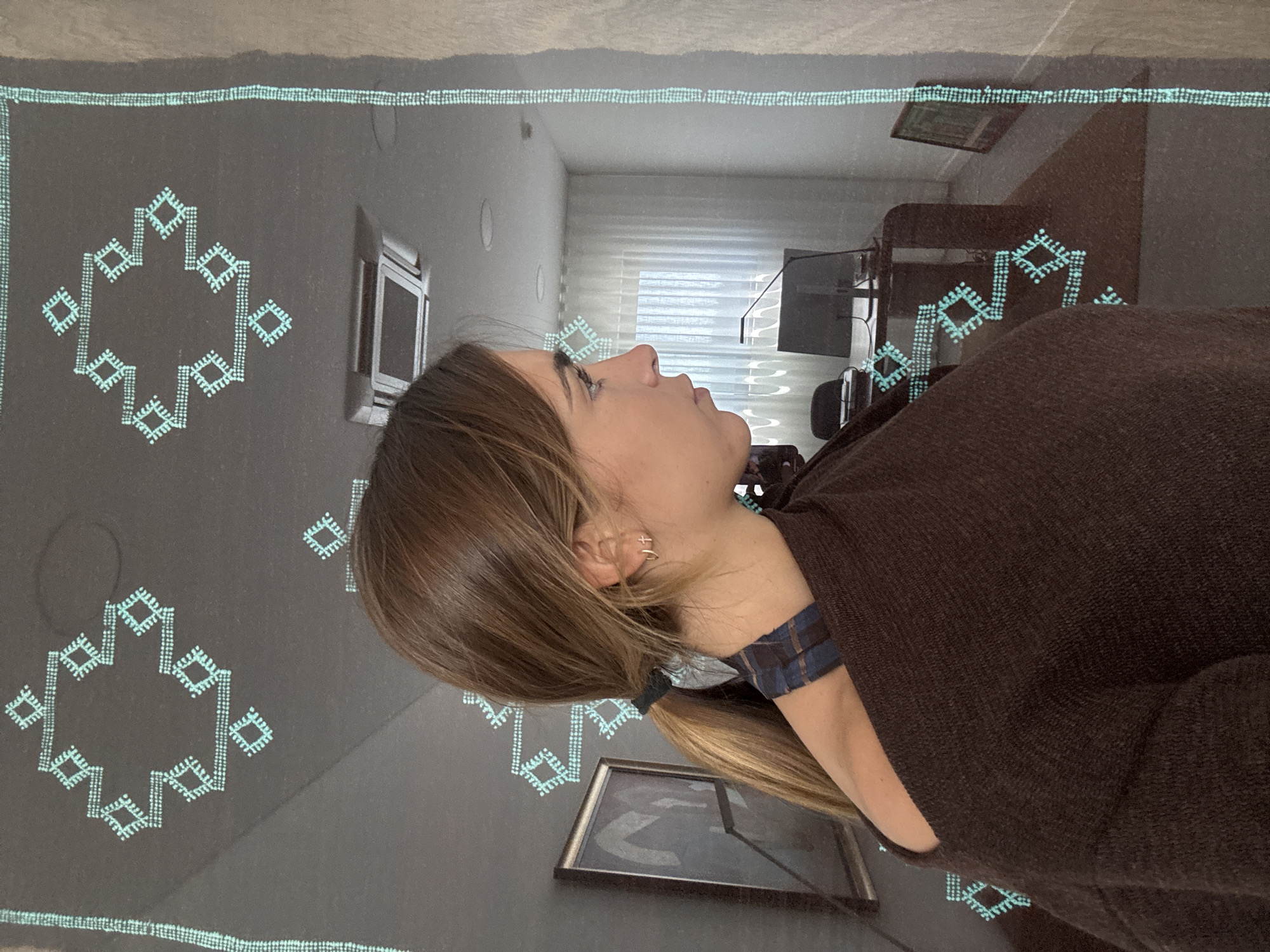 The image shows a person with brown hair, wearing a brown sweater and a patterned neck covering. They are indoors, possibly in a room with a decorative rug. The rug is patterned with geometric designs. The background shows a glimpse of an interior room setting with a window, desk, and artwork. The image does not directly indicate the location being Torres Vedras, Portugal.