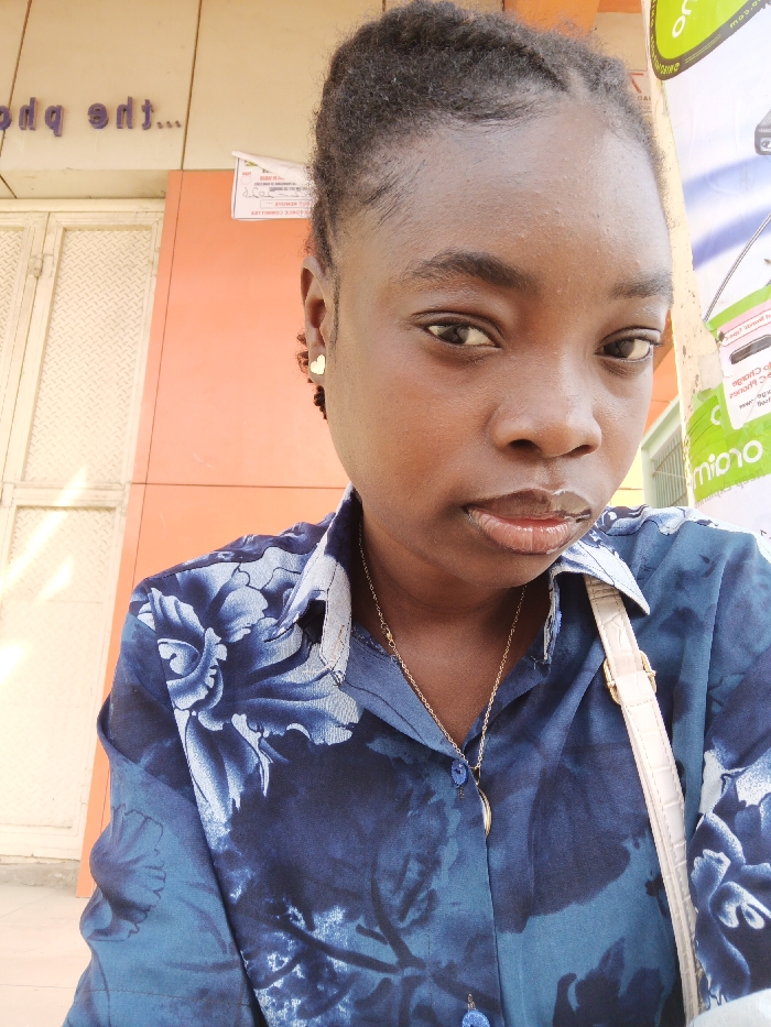 A medium-shot portrait depicts a dark-skinned individual, identified as a young adult female, looking directly at the camera with a neutral expression. Her dark, textured hair is styled back. She wears a dark blue collared shirt featuring a lighter blue floral pattern and a small, gold-colored heart-shaped stud earring in her visible right ear. A thin gold-colored chain necklace with a curved, light-colored pendant is visible, along with the light-colored strap of a shoulder bag crossing her right shoulder. The background to the left features an orange-colored wall segment positioned above a light-colored, diamond-patterned metal shutter or door. Above the orange wall, an inverted purple sign partially displays text such as "tdq eht...". To the right, a vertical pole or column is partially covered with promotional posters, including a prominent green section with white text, where "orio" is discernible. The scene is located in the city of Fagge A, Nigeria.