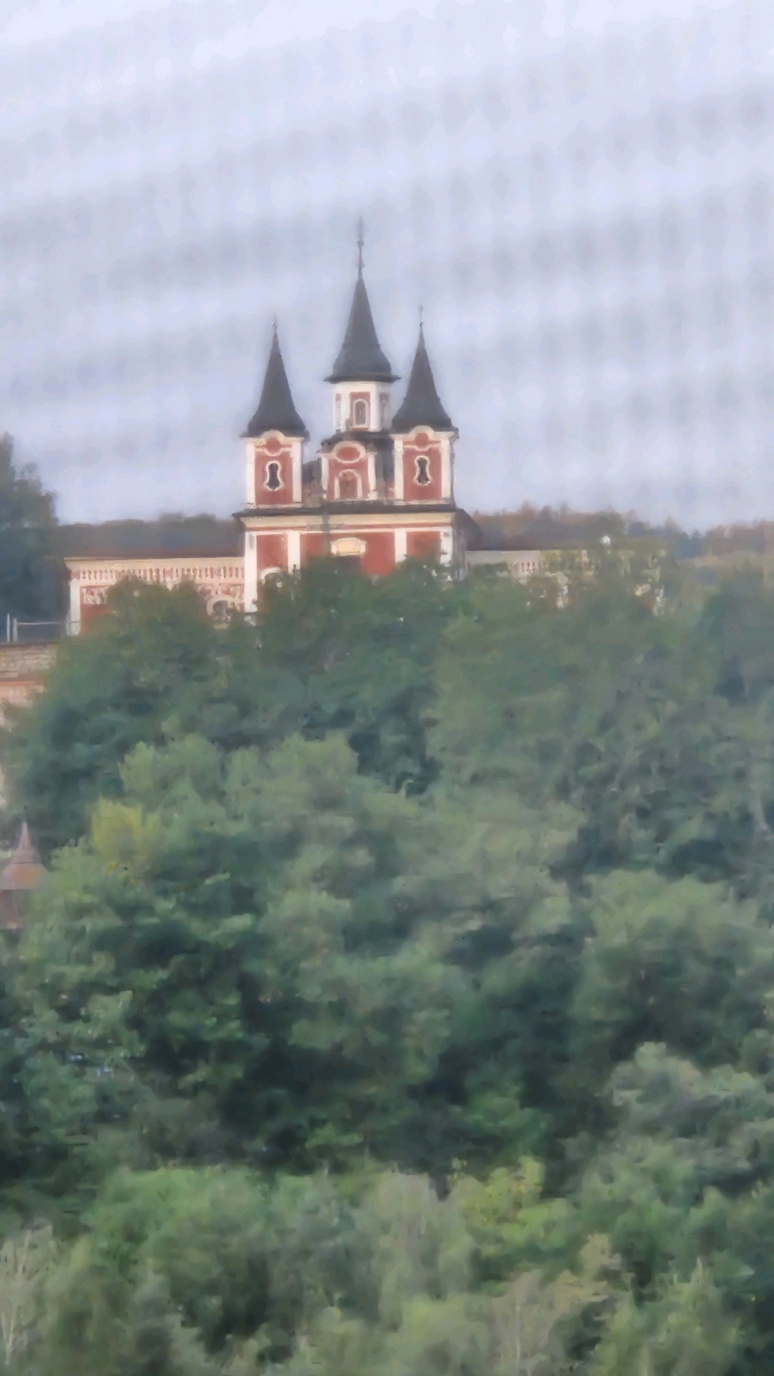 The image depicts a religious building, likely a church or monastery, with a distinctive red and white facade and three prominent spires. The structure is situated on a slight incline, surrounded by lush green trees and foliage. The sky is overcast, giving the scene a muted, diffused light. The architectural style suggests it might be of historical significance. This building is identifiable as the Kláštor svätej Alžbety (St. Elizabeth's Monastery) in Prešov, Slovakia. The monastery complex is a notable landmark within the city.