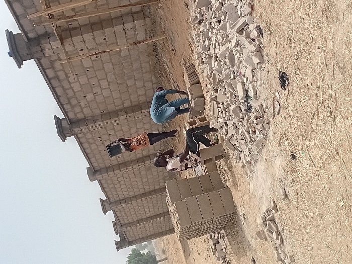 The image displays an outdoor construction scene in Ayama, Nigeria. Three adult males are present near a partially constructed perimeter wall made of light grey concrete blocks. The wall extends across the mid-ground and features multiple vertical pillar sections topped with decorative capstones.
In the left foreground, a male individual is seated on a stack of unlaid concrete blocks. He is wearing a light-colored, patterned shirt and dark trousers. His right arm is bent and resting.
In the center-right, another male, dressed in an orange t-shirt with visible text "500" and dark trousers, is standing and holding a dark bucket with both hands, lifted above his head, suggesting material transport.
To the far right, a third male, clad in a light blue long-sleeved garment, is bent over, engaged in ground-level work, possibly arranging or handling construction materials.
A large pile of broken concrete blocks and rubble occupies the central foreground. A distinct stack of whole concrete blocks is located in the lower-left. The ground is dry, light brown earth with sparse dry vegetation. In the far right, two rudimentary wooden poles, possibly temporary supports, stand vertically. The background on the left shows distant green trees under a hazy, bright sky.