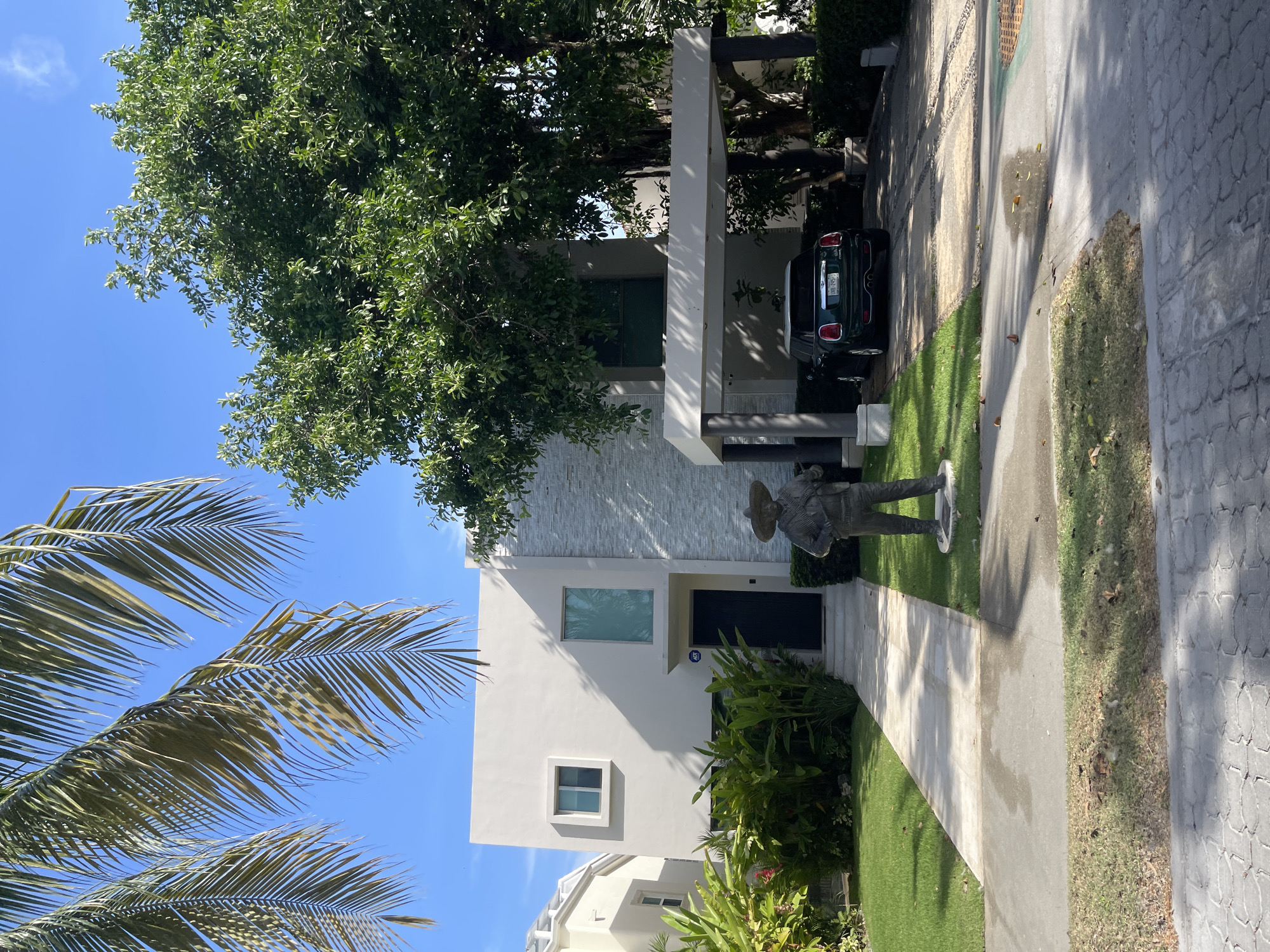 The image shows a modern two-story house with a white exterior and gray stone accents, located in Playa del Carmen, Mexico. The house has a green front door and a covered carport with a dark green car parked underneath. A statue of a man in a sombrero stands on the lawn in front of the house. Lush tropical foliage surrounds the property, including palm trees visible in the foreground. The sky is clear and blue. A paved walkway leads to the front door, and a concrete driveway extends to the street. Another house is partially visible on the left of the frame.