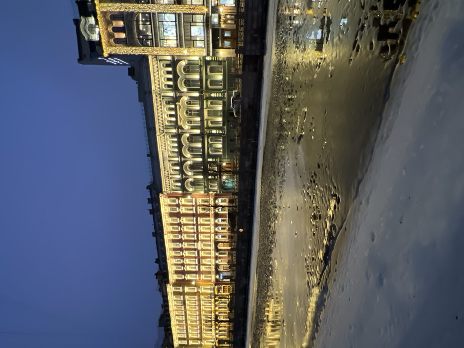 The image displays a nighttime winter scene along the Griboyedov Canal Embankment in Saint Petersburg, Russia. The sky above is dark blue. In the immediate foreground, a snow-covered bank slopes towards the canal. The canal itself is partially frozen, featuring a mixture of dark open water and irregular patches of ice. The water's surface reflects the yellow and white artificial lights from the buildings on the opposite bank.
Across the canal, a stone embankment wall is visible, bordering a paved road. A single light-colored vehicle is parked on this road. Bare, deciduous trees are sparsely distributed along the embankment. A continuous row of five multi-story buildings, characteristic of 19th and early 20th-century Russian architecture, forms the background. All buildings are brightly illuminated with exterior accent lighting and visible interior lights.
From left to right:
1. A five-story building with a light yellow-orange facade and numerous uniformly lit windows.
2. An adjacent five-story building with a reddish-orange facade and similar window illumination.
3. A six-story building with a green-grey facade, featuring prominent arched windows on its upper floors and decorative architectural details, all brightly lit.
4. The tallest structure on the far right is a multi-story building, identified as Singer House (Dom Knigi). Its darker facade is extensively adorned with vertical strings of yellow festive lights, and its multiple large windows are brightly illuminated. The building is topped with a distinctive ornate structure.
No individuals or explicit actions are discernible within the frame. The scene conveys a cold, urban winter evening.