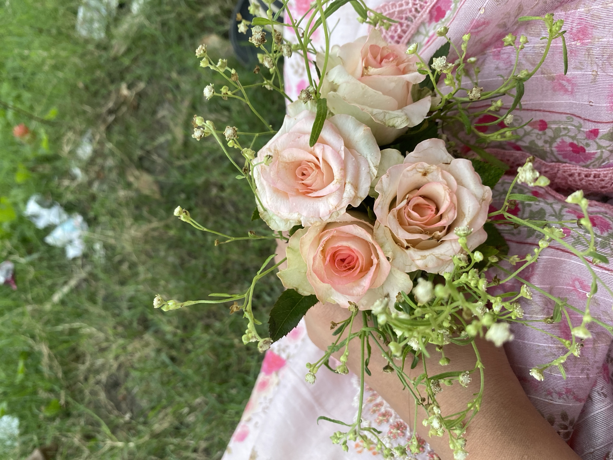In Tarabo, Bangladesh, a person's forearm rests on a pink fabric with a floral pattern, holding a small bouquet of pale pink roses and delicate white wildflowers. The background consists of green grass with some scattered debris, suggesting an outdoor setting, possibly a park or garden. The lighting appears to be natural daylight, casting soft shadows.