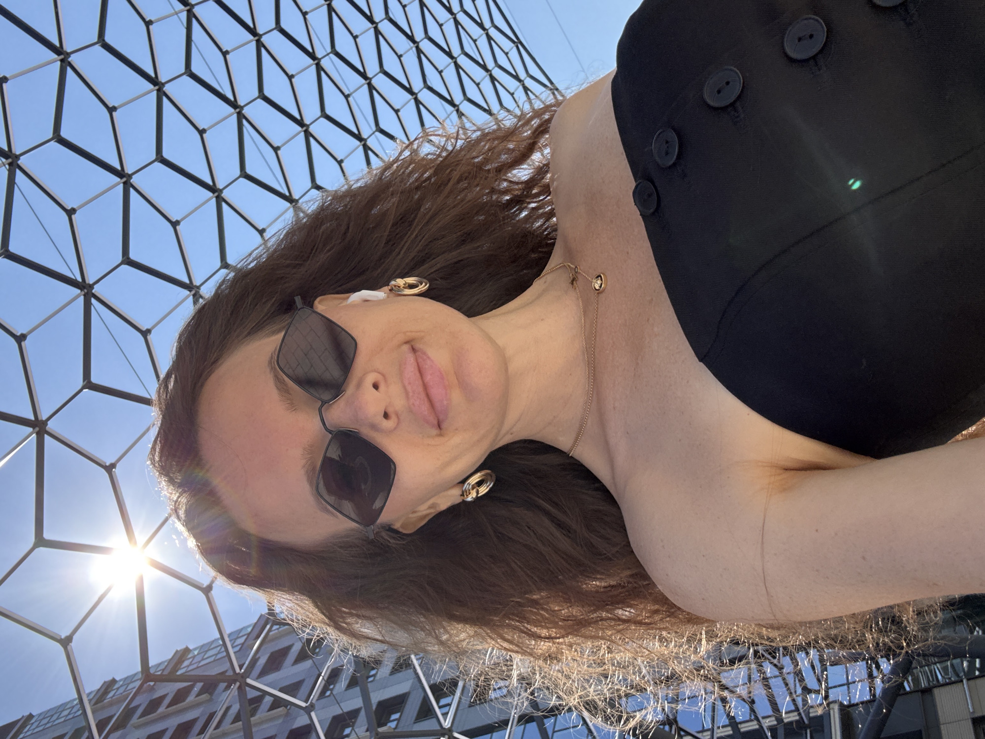 A woman takes a selfie outdoors, with a modern building and a geometric canopy overhead. She is wearing sunglasses, hoop earrings, a necklace with a small circular pendant, and a black off-the-shoulder top with four visible buttons. The sun is shining brightly, creating lens flare. The image is taken from a low angle, looking up at the woman and the sky. The woman is likely in a city, possibly Moscow, Russia, given the contemporary architecture and urban setting suggested by the building and canopy structure.
