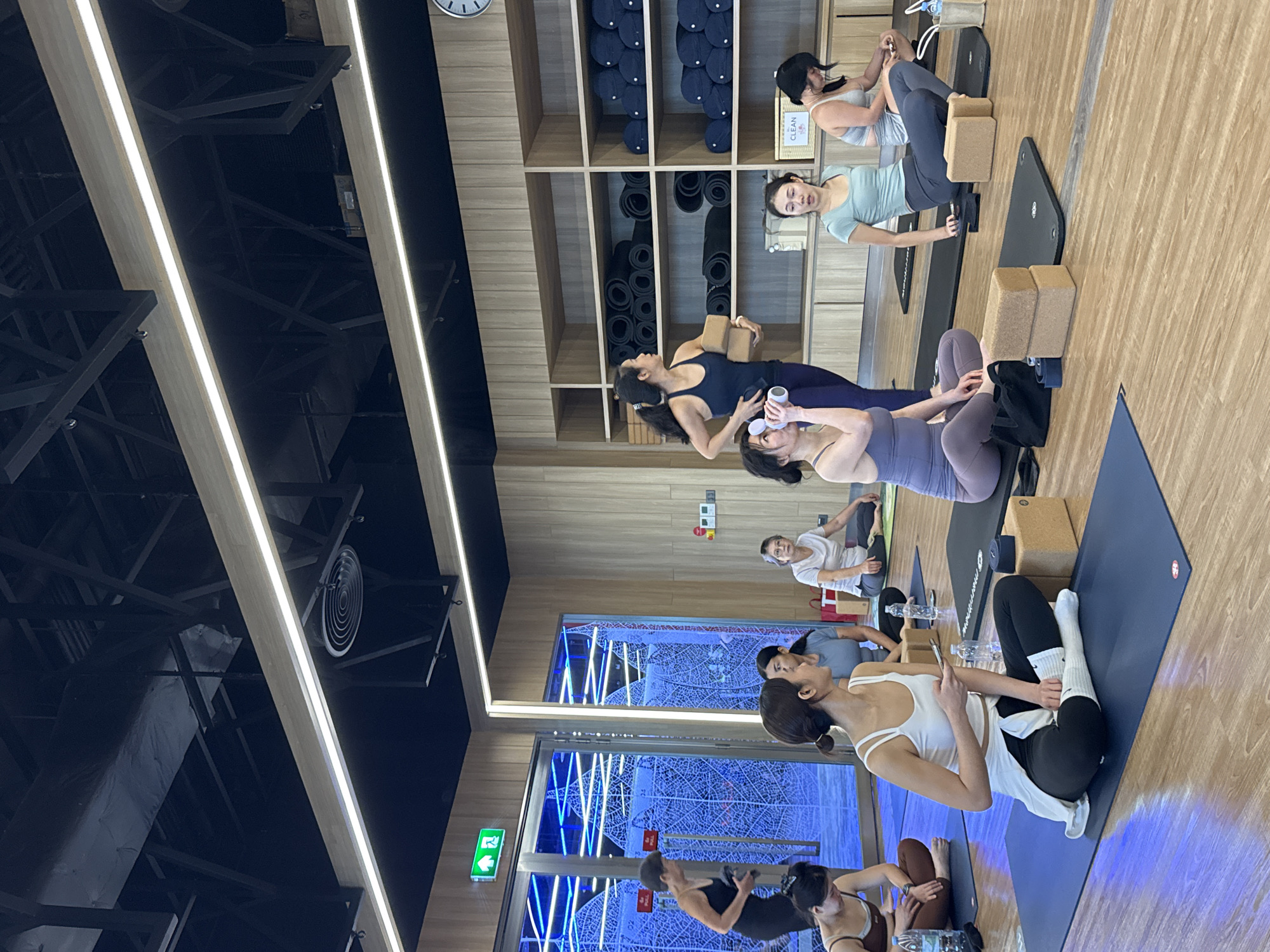 A yoga class is in session at a modern studio located in Nong Pa Khrang, Thailand. The room has light wood-paneled walls, a wooden floor, and a dark ceiling with industrial lighting. In the foreground, several women are seated on blue yoga mats, some in meditative poses. One woman in the center foreground wears a white tank top and black leggings, holding a phone. To her left, a woman in a purple tank top and leggings drinks from a water bottle. To her right, a woman in a teal top and grey leggings looks ahead, with two yoga blocks in front of her. In the background, a woman in a dark blue tank top and purple leggings stands holding two yoga blocks, facing a shelf unit stocked with rolled yoga mats and dark blue bolsters. Another woman in a white t-shirt and grey pants sits further back on the right. To the left, a woman in a black tank top and leggings walks past a glass door with blue neon lighting and a decorative frosted pattern. The overall atmosphere is calm and focused.