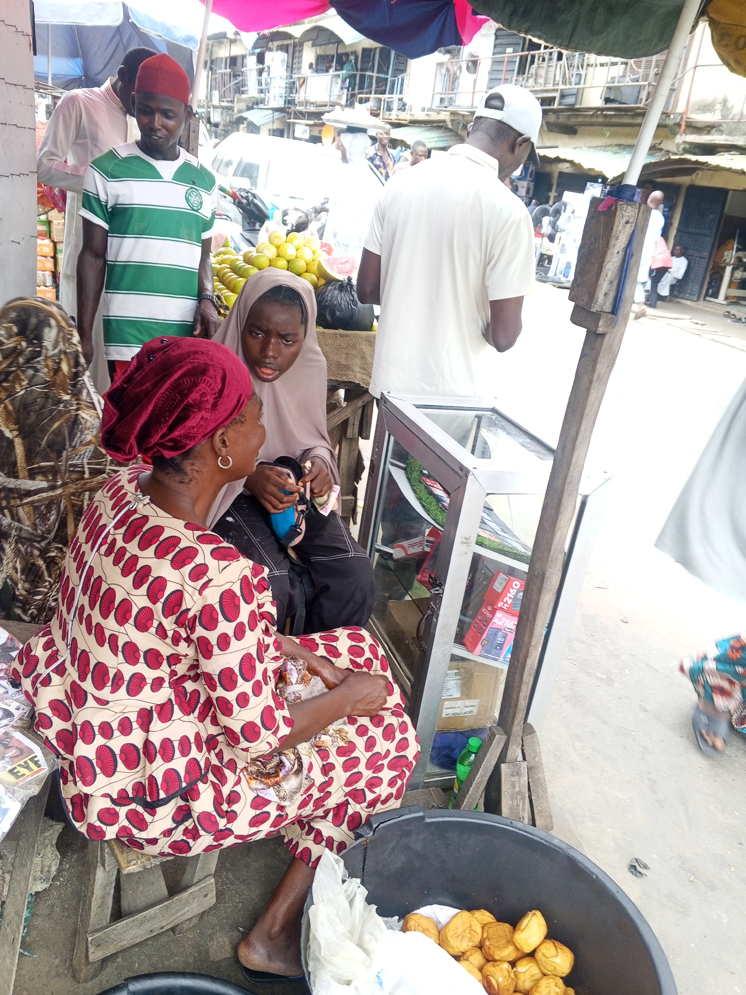 In the city of Okene, Nigeria, a bustling market scene unfolds. In the foreground, a woman in a vibrantly patterned dress and a deep red headscarf sits on a stool, her gaze directed towards a young girl beside her. The girl, wearing a light brown hijab, holds a small blue object, possibly a phone case or a wallet, while her mouth is slightly open as if speaking. Behind them, a man in a green and white striped shirt and a red cap stands, looking down. To the right, a glass display case exhibits various items, including boxed electronics. The market environment is evident with awnings overhead and other individuals visible in the background, some tending to stalls and others walking through the crowded area. A large black bin filled with golden-brown fried pastries sits prominently in the lower right corner of the frame, suggesting food sales are also taking place. The ground is paved and appears somewhat dusty, typical of an outdoor market.