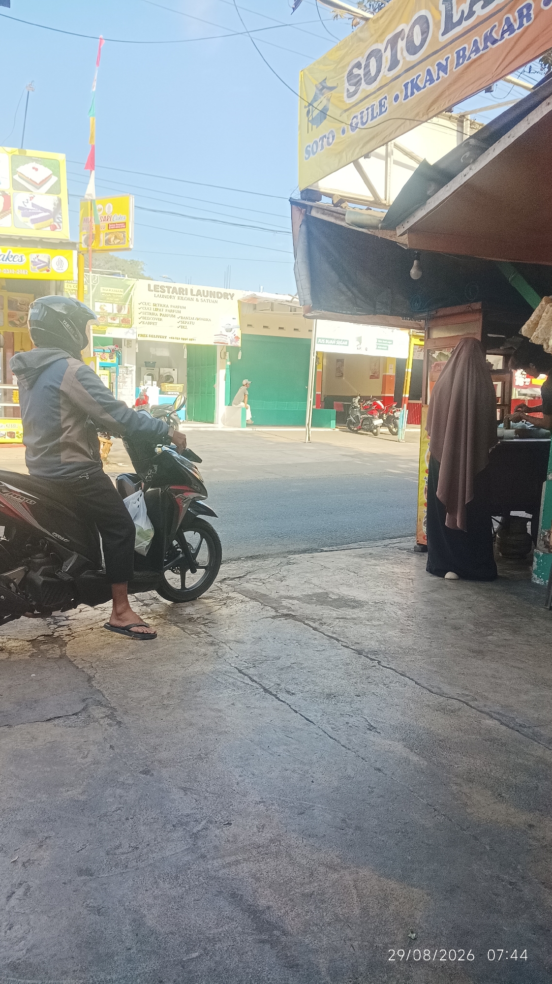 A full outdoor shot shows a street scene with several businesses and pedestrians in Tasikmalaya, Indonesia. The day is bright and sunny. In the foreground, a person on a motorcycle faces to the left. The motorcycle is red and black. The person is wearing a gray hooded jacket, black pants, and black flip-flops. A white plastic bag hangs from the handlebars. The pavement is cracked and wet in places.
In the middle ground, a building with a yellow banner featuring the words "SOTO LA..." is visible. Below this banner, there are signs for "LESTARI LAUNDRY" and "SAPI Cakes". A person wearing a brown hijab and a black outfit stands to the right, facing away from the camera. Another person is partially visible behind a counter on the far right. Two red motorcycles are parked in front of a green building in the background. A person sits on a concrete ledge in front of the green building.
The time stamp "29/08/2026 07:44" is visible in the bottom right corner of the image.