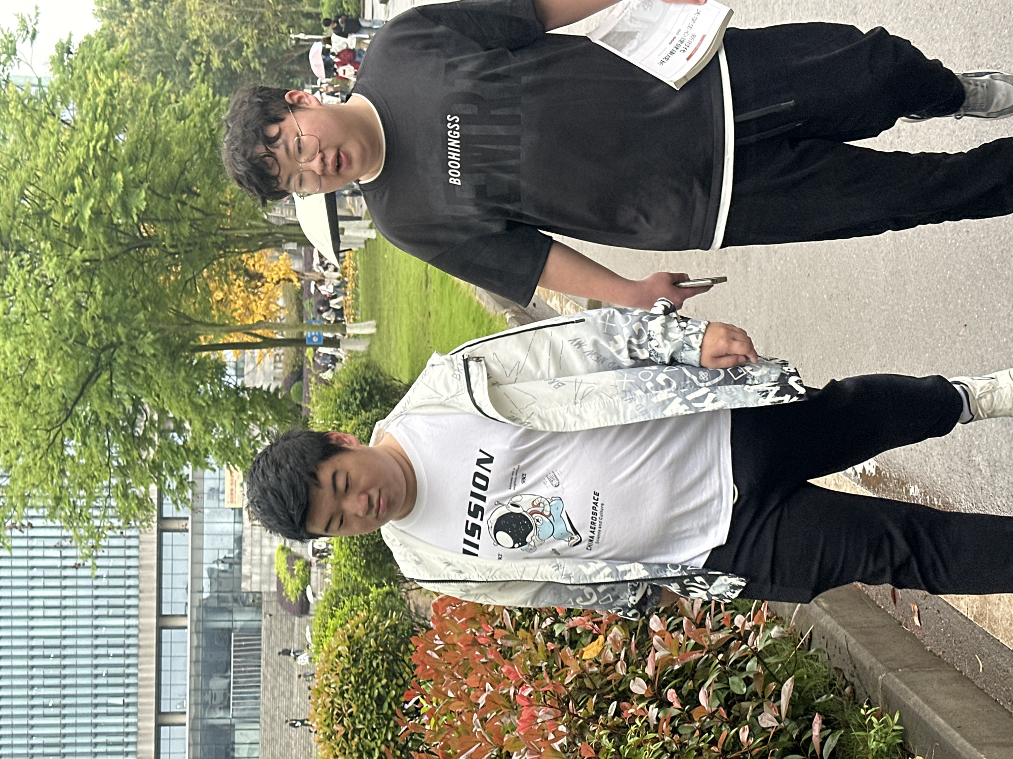Two Asian men are standing on a sidewalk near a building. The man in the foreground is wearing a white t-shirt with an astronaut graphic and the words "MISSION" and "CHINA AEROSPACE" on it, a light gray jacket, and black pants. The man in the background is wearing a black t-shirt with the word "BOOHINGSS" on it, black pants, and glasses, and he is holding a book with Chinese characters on it. The location is Jiangjin, China, as indicated by the Chinese writing on the book.