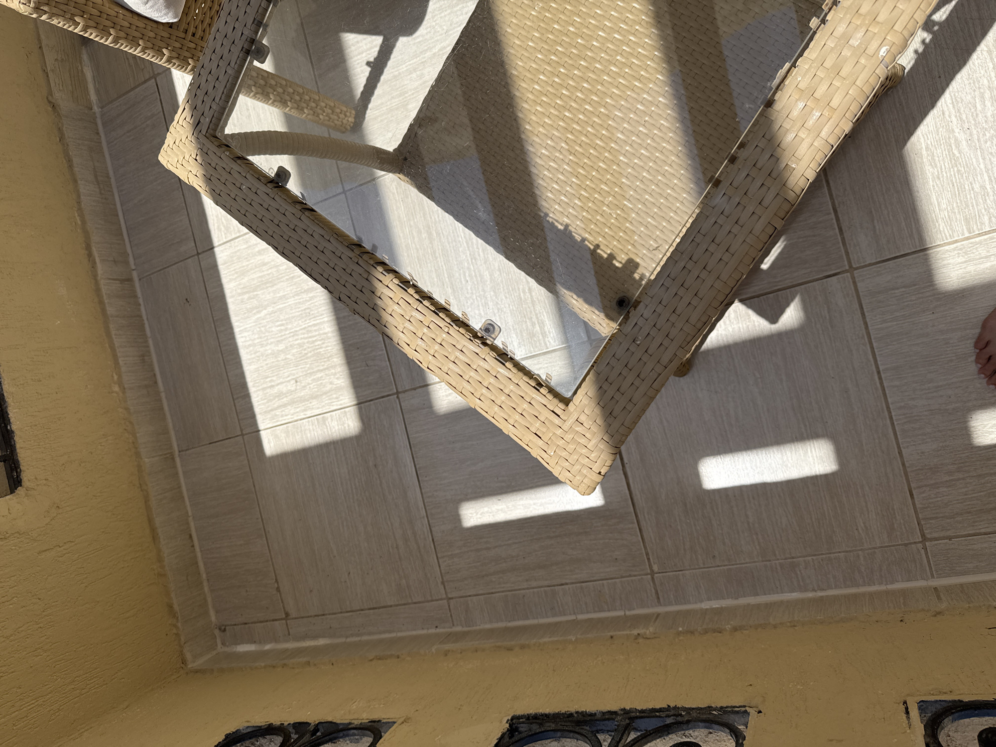A top-down, close-up shot captures a corner of an outdoor tiled patio, bathed in dappled sunlight casting distinct rectangular shadows. A wicker-style table with a glass top occupies the right side of the frame, its woven texture visible. The wall on the left is painted a textured yellow and features decorative, dark-rimmed circular inlays. The tiled floor is composed of square, light-colored tiles. A sliver of a foot, bare and with painted toenails, is visible at the bottom center of the frame, suggesting a person is present but not fully in view. The scene appears to be on a balcony or patio, possibly in a warm climate given the sunlight.