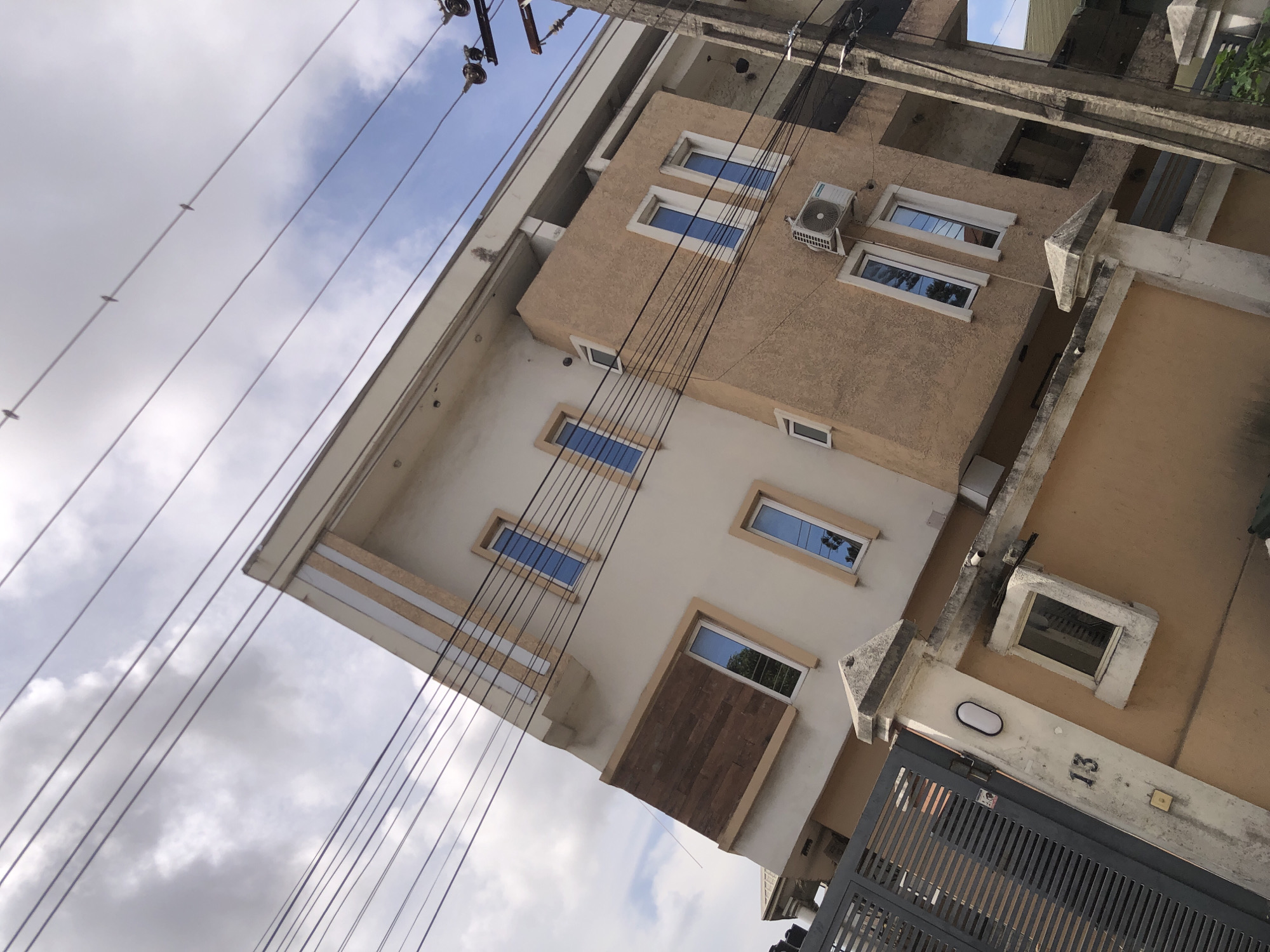 This image captures an upward-angled, oblique view of a multi-story building, likely residential, against a partly cloudy sky. The building features a light tan-colored upper facade and a lighter, off-white lower section, with prominent white-framed rectangular windows, some reflecting the blue sky. An outdoor air conditioning unit is mounted on the tan wall of the upper floor.
Numerous electrical wires stretch diagonally across the frame, connected to utility poles, with parts of the poles visible at the top. Several small birds are perched on these wires. The sky displays a mix of bright blue patches and white, wispy clouds.
Architectural details include concrete ledges, beams, and decorative elements. Towards the bottom right, a dark, slatted metal gate is partially visible. On the concrete wall near the gate, the number "13" is clearly displayed, indicating the address. An oval-shaped outdoor light fixture is also mounted on this wall.
The scene depicts a static urban or suburban environment during daytime, under partly cloudy weather conditions, in Ado, Nigeria. No human activity is visible.