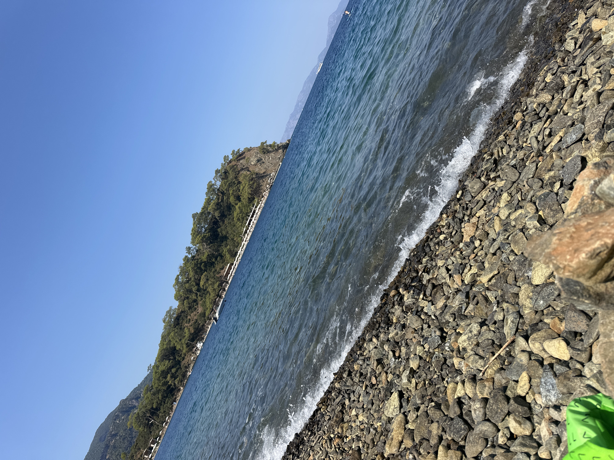A diagonal shot captures a serene beach scene along the coast of Fethiye, Turkey. The foreground is dominated by a pebbled shoreline, with small waves gently lapping at the shore, creating white foam and ripples. Beyond the water, a lush green hillside covered with trees slopes down towards the sea. The hillside is dotted with small white structures, possibly beach umbrellas or cabanas, suggesting a resort or tourist area. A steep, rocky outcrop juts out into the water on the right side of the frame, also covered in vegetation. The sea is a deep blue, transitioning to lighter turquoise near the shore. In the distance, hazy mountains form the backdrop. The sky is a clear, cloudless blue. No people are clearly visible in the image, only the environment.