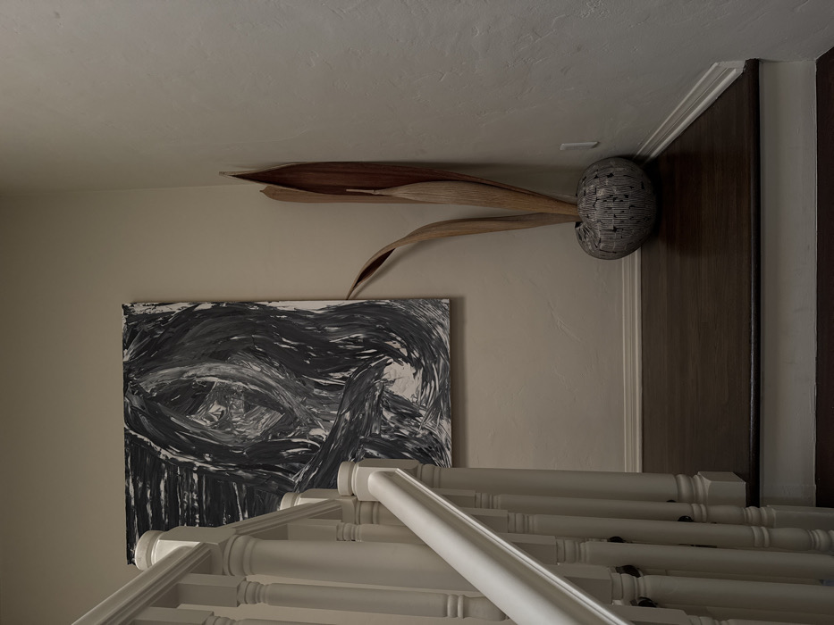 This image depicts an indoor residential stairway landing or hallway. A white wooden staircase railing with turned balusters ascends from the lower-left foreground. On the light-colored, textured wall to the left, a large, unframed rectangular abstract painting hangs, featuring a composition of black and white swirling brushstrokes. In the bottom right corner of the room, a spherical, textured vase with a striped or grooved pattern in muted grey and brown holds several tall, dried, naturally curled brown plant fronds. The floor is covered with dark brown wooden planks, bordered by white baseboards. A white electrical outlet or light switch plate is visible on the wall to the right of the vase. The specific location within Los Angeles, United States of America, cannot be determined from the visible elements. No individuals, specific actions, or interactions are present in the scene.