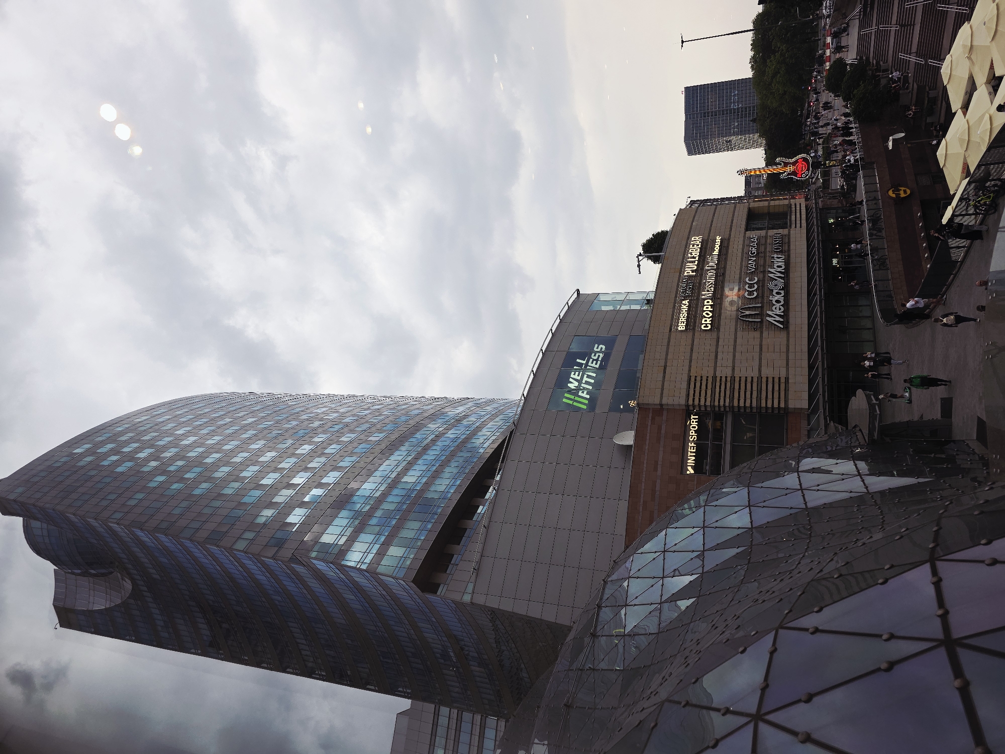 This eye-level shot depicts a contemporary urban landscape in Warsaw, Poland. Prominently featured is a modern high-rise building with a curved design and glass facade. Adjacent to it, a multistory retail complex hosts a variety of stores like Bershka, Pull & Bear, Cropp, Massimo Dutti, CCC, Media Markt, Oysho, New Fitness, and Inter Sport. Shoppers are seen on the sidewalk in front of the retail area. In the background, there are a few pedestrians, some trees, a large building and a Hard Rock Cafe sign. Overhead, the sky is overcast with soft, diffused light.