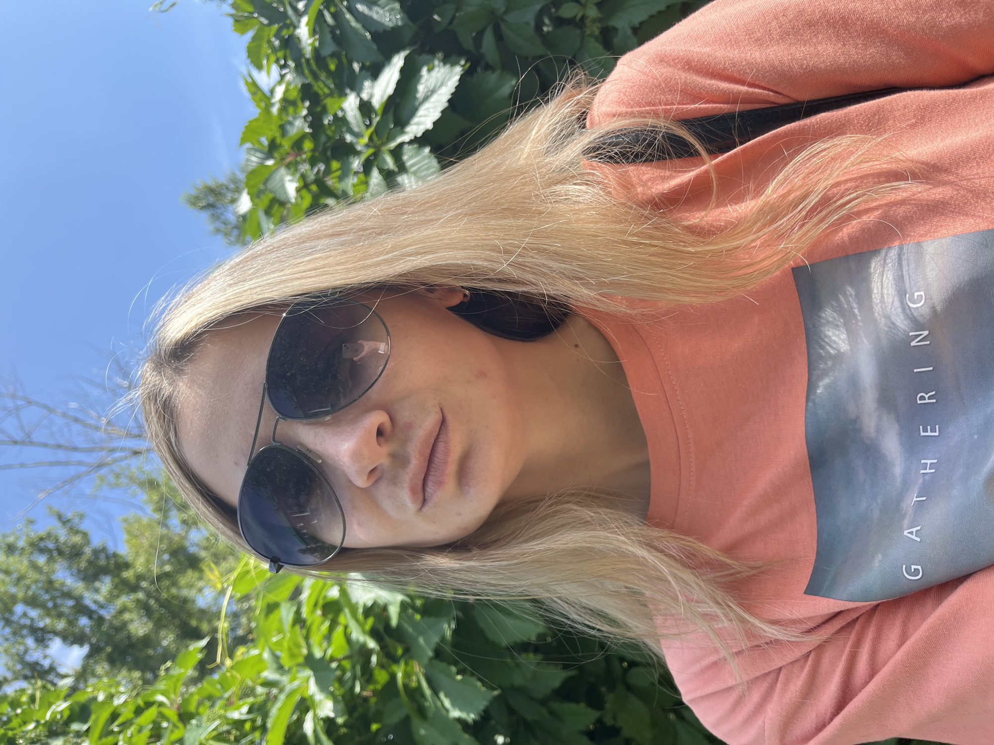 A young woman with blonde hair and sunglasses is taking a selfie outdoors. She is wearing a peach-colored t-shirt with the word "GATHERING" printed on it. Behind her, there is lush green foliage and a bright blue sky. The image appears to have been taken in Chaikovsky, Russia, likely in a park or garden, given the natural surroundings.