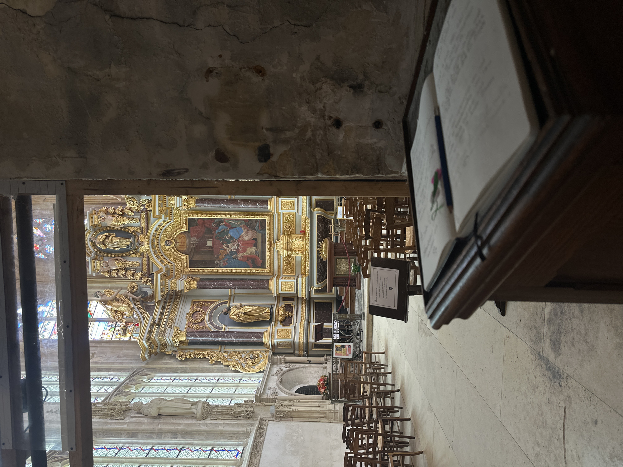 The photograph depicts the interior of a church, likely Saint-Jacques in Dieppe, France, given the prominent ornate altar and stained-glass windows. In the foreground, a wooden lectern holds an open book with a pencil and a small colorful ribbon marking a page. The book appears to be filled with handwritten text. To the left, rows of empty wooden chairs with woven seats are arranged on a tiled floor. Beyond the chairs, large stained-glass windows with intricate tracery are visible. The focal point of the image is the highly decorative altar, featuring a large painting above, gilded sculptures, and marble columns. A statue of a man in gold robes stands in a niche above the altar. The wall on the right side of the frame is rough and cracked, contrasting with the refined architecture of the church interior. The lighting suggests an overcast day or indirect sunlight. No individuals are present in the scene.