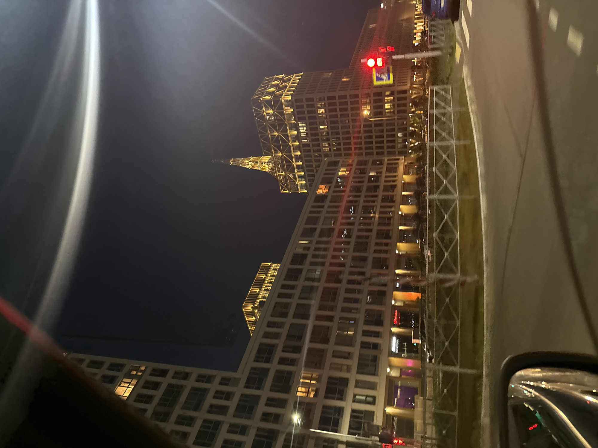 The image depicts a nighttime view of a modern architectural complex in Saint Petersburg, Russia. The foreground shows a curved road with a pedestrian crossing and a safety barrier. A car's side mirror is partially visible on the left, reflecting city lights and the night sky.
In the mid-ground, several multi-story buildings are illuminated from within and by exterior lighting. The buildings have a repetitive facade design with numerous windows. One prominent building on the right is taller and features a distinctive illuminated spire and a geometric lattice structure at its top. A traffic light showing red is visible on the right side of the road. The sky is dark, indicating nighttime, with a faint, bright light source creating a halo effect in the upper right quadrant of the image. The overall impression is one of a contemporary urban environment at night.