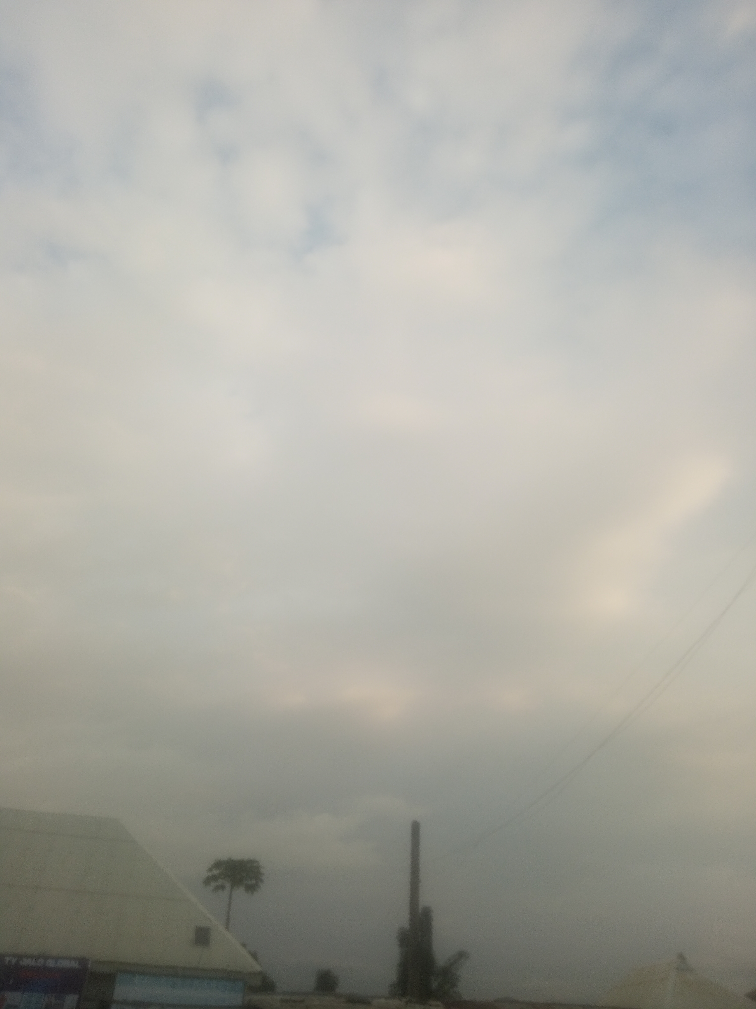 The image captures a cloudy sky above the city of Serti, Nigeria. In the foreground, a portion of a white, angled building is visible on the left, with some signage that is partially obscured. To the right of the building, a tall, thin smokestack or industrial structure stands against the hazy sky. Further to the right and slightly behind the smokestack, a solitary palm tree is silhouetted against the overcast backdrop. A faint power line can be seen stretching diagonally across the upper right portion of the frame. The overall atmosphere is subdued due to the heavy cloud cover.