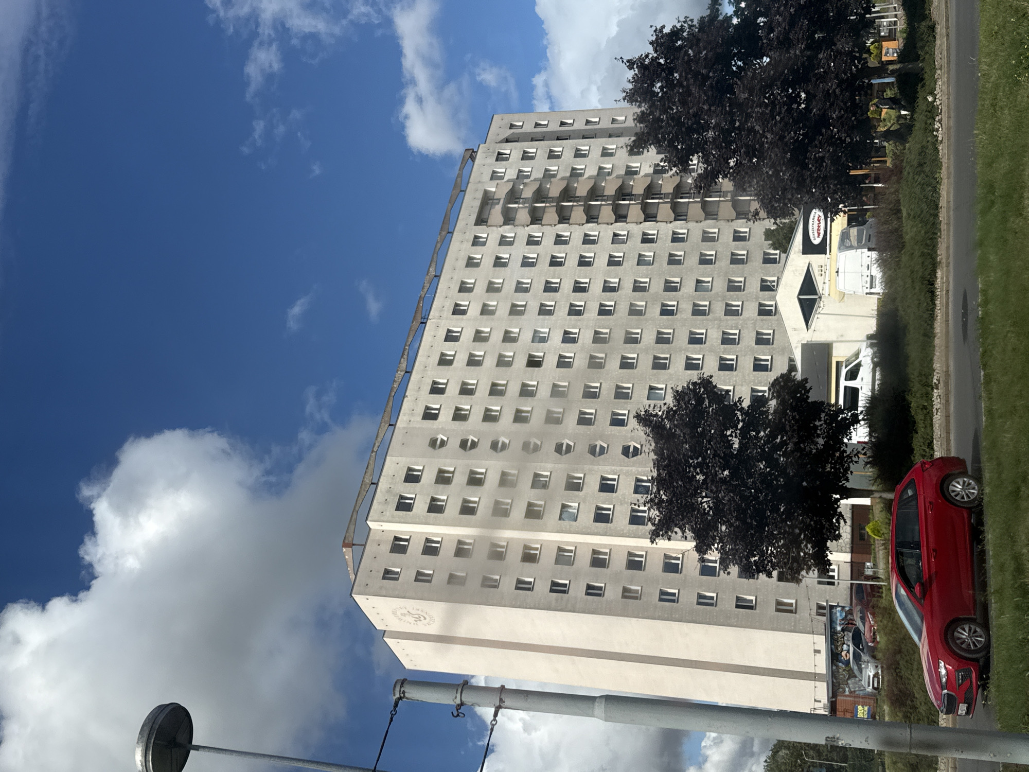 The image shows a tall, light-colored apartment building in Szczecin, Poland, under a partly cloudy blue sky. A street lamp is visible on the left side of the frame, as is a red Skoda station wagon driving on a road in front of the building. There are several trees with dark foliage on the right side of the building, and a low-rise building with a sign that reads "SZASZELKARMA MERKURY" is visible between the apartment building and the trees. There are a few parked cars in front of the low-rise building. The apartment building has many windows and a balcony structure on its side. A circular emblem with text is present on the upper left facade of the building.