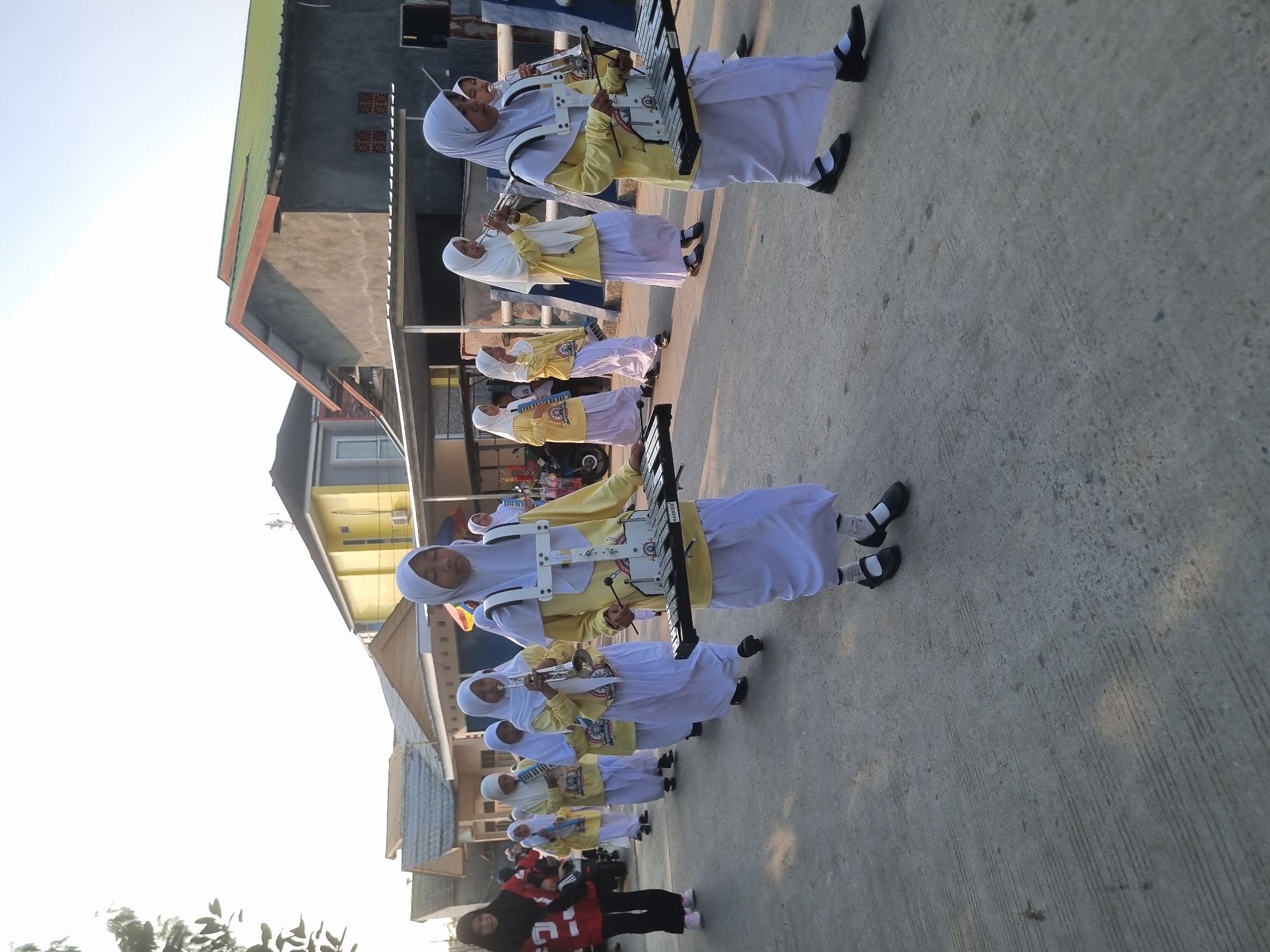 The image depicts a group of young girls in a marching band, dressed in yellow tops and white skirts and hijabs, parading down a paved street in what appears to be a residential area. They are carrying musical instruments, including what look like glockenspiels and trumpets. The girls are walking in a line, some of them playing their instruments. In the background, there are houses with various architectural styles, some with green roofs. The sky is bright, suggesting it is daytime, possibly late afternoon due to the warm light. A person in a red jacket and black pants is walking on the left side of the street, slightly ahead of the marching band. There are also motorcycles visible on the left side of the image. The street is made of concrete and shows some signs of wear and tear. The overall scene suggests a local community event or a school activity in Samarinda, Indonesia.