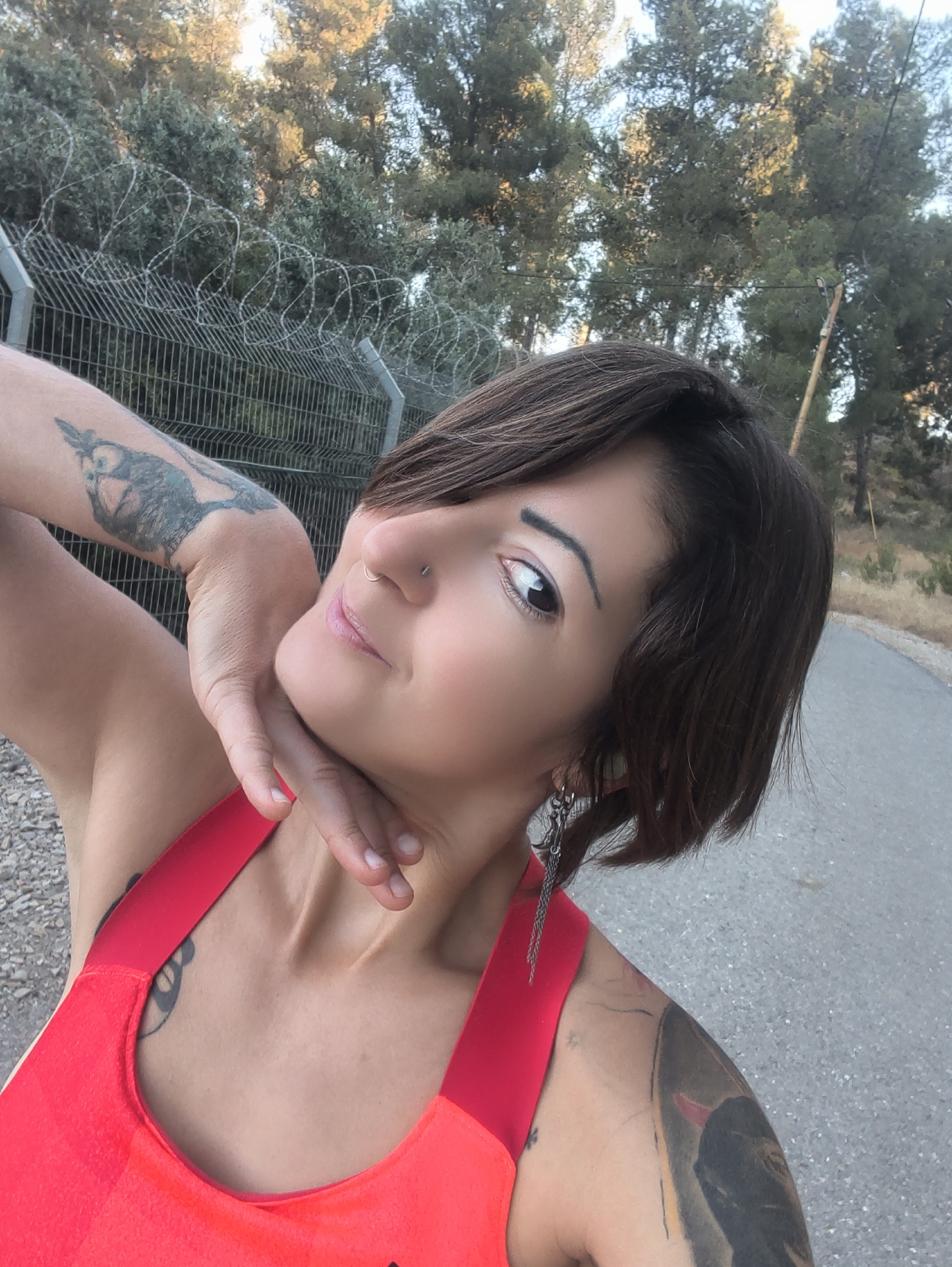 In a daylight outdoor scene, likely in the Lachish Regional Council, Israel, a woman with short, dark hair and tattoos on her arms and shoulder poses for a photo. She is wearing a bright red sleeveless top. Her left arm is raised, with her hand resting on her neck. She has a nose ring and is looking towards the camera with a slight smile. Behind her, a chain-link fence topped with barbed wire separates the area from a wooded hillside with trees. A paved road runs along the right side of the frame.