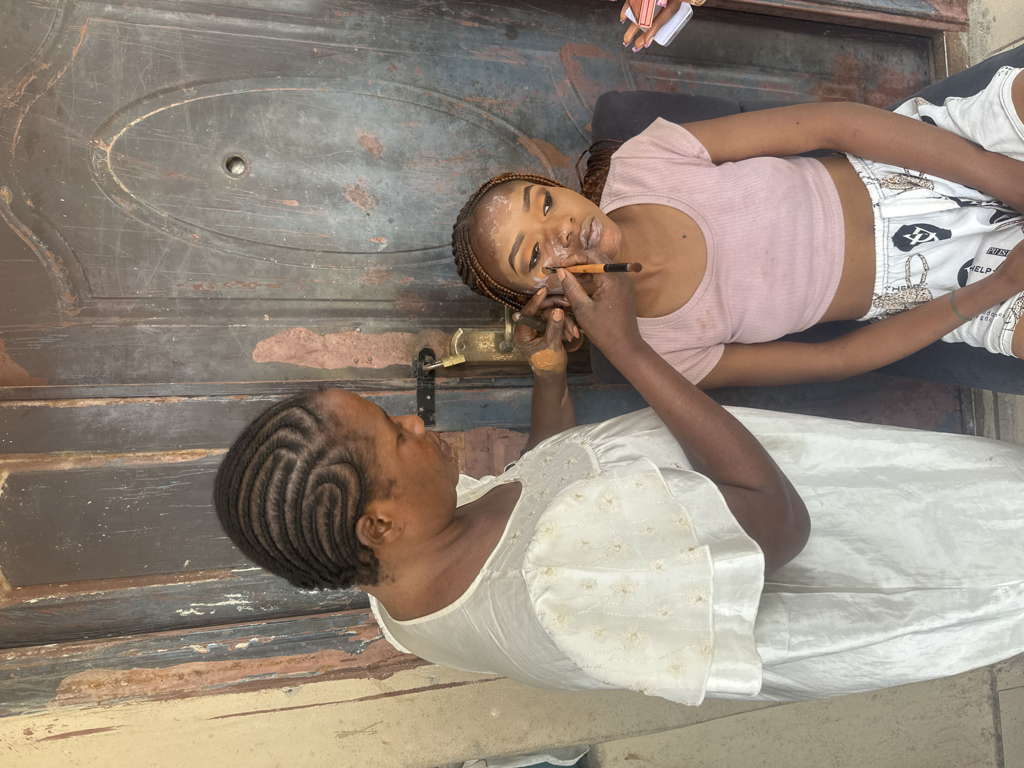 In Badagry, Nigeria, a woman with braided hair is applying makeup to another woman seated in front of a rustic, weathered brown door. The woman applying makeup wears a white dress with decorative embroidery on the sleeves and neckline. The seated woman, with her hair in braids, is wearing a pink crop top and patterned shorts, and appears to be in the process of having her makeup done, with foundation and eye shadow already applied. The background consists of a textured, aged door with visible paint chips and a lock mechanism.