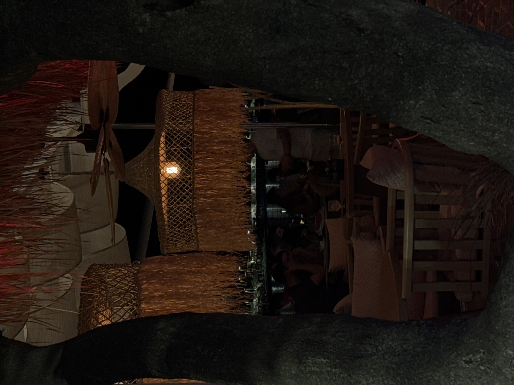 The image depicts an outdoor seating area at a establishment in Petrovac centar, Montenegro, likely a restaurant or bar, as suggested by the presence of tables, chairs, and patrons. The scene is dimly lit, creating an atmospheric ambiance, possibly during evening hours.

Dominating the foreground are the dark, textured trunks of two large trees, framing the view into the establishment. Above, woven bamboo light fixtures with a fringed, straw-like material hang, illuminating the space with a warm glow from visible bulbs. One fixture, in the center of the image, has a single incandescent bulb. A ceiling fan is visible in the background, suggesting a warm climate.

In the mid-ground, several people are seated at tables, some appearing to be in conversation. The furniture consists of light-colored wooden chairs and tables, with comfortable-looking cushions on some of the seating. The background shows more of the interior or covered seating area, with additional lighting and possibly a bar. The overall impression is of a relaxed, social setting.