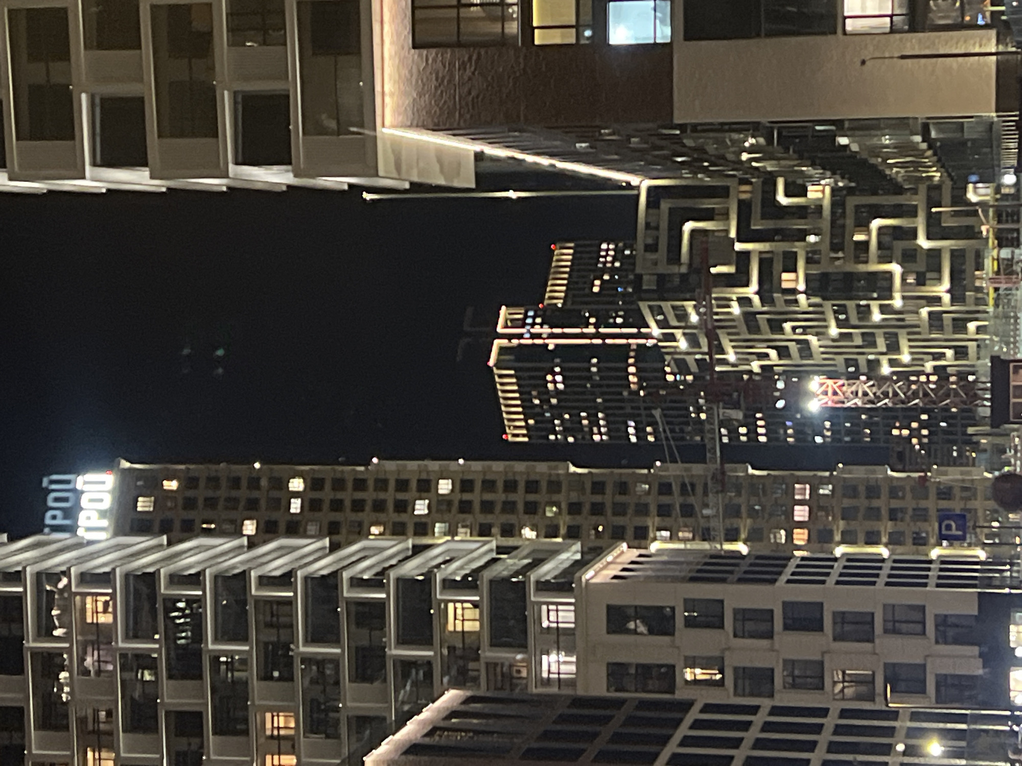 The image depicts a nighttime urban landscape in Moscow, Russia. The view is dominated by a mix of modern architectural styles. On the left and bottom are buildings with a blocky, geometric design. A central high-rise building displays an intricate facade design illuminated with many lights. The skyline is illuminated with numerous windows and other building lights, and what appears to be construction cranes and signage are visible in the cityscape.