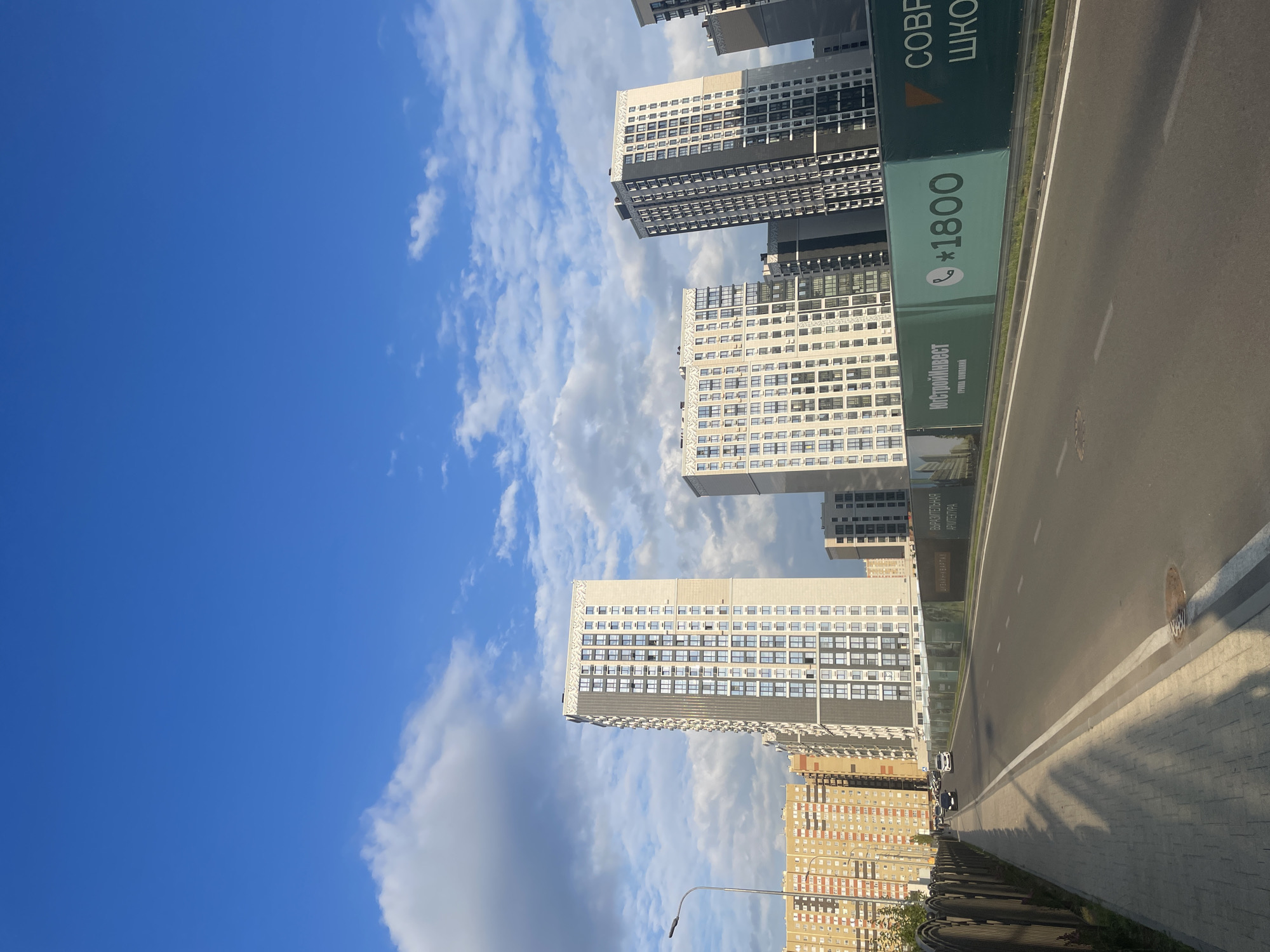 This image captures a daytime scene in Stavropol, Russia, featuring a street lined with modern apartment buildings. The sky is blue with scattered white clouds. On the left side of the road, a paved sidewalk runs alongside a dark metal fence. The road itself is asphalt with white lane markings. A row of green construction hoardings runs along the right side of the road, displaying advertisements for a company with a phone number and text in Cyrillic. Several cars are visible driving on the road, and pedestrian traffic is not apparent. The buildings are tall, multi-story structures with numerous windows, indicating a residential or mixed-use urban development. The overall impression is that of a developing area within the city.