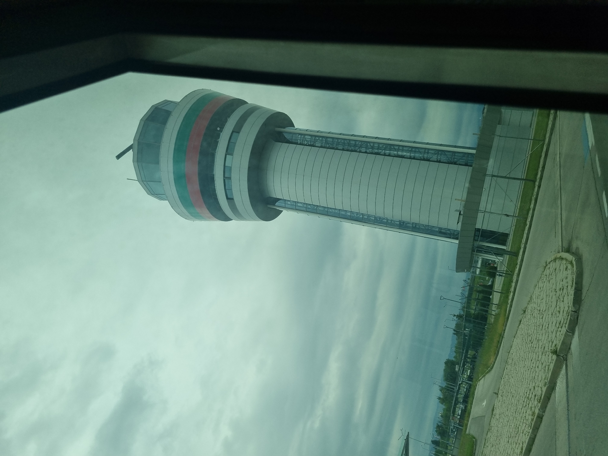 The image depicts the Sofia Airport Control Tower, located in Sofia, Bulgaria. The tower is tall and cylindrical, with a distinctive top section colored in white, green, red, and black stripes. It is connected to a lower building via an enclosed walkway. The scene is viewed from inside a vehicle, framed by a window. Outside, there is a paved area, grass, and trees. The sky is cloudy. There are no people visible in the image.
