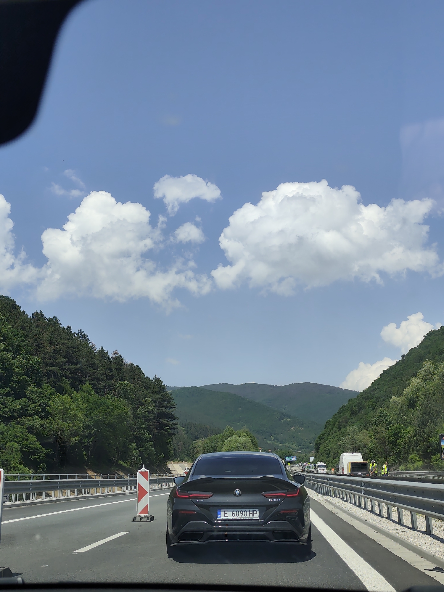 The image depicts a highway scene on a bright, sunny day, with dense green mountains flanking both sides of the road under a blue sky with scattered white clouds. The perspective is from inside a vehicle, evidenced by a dark, curved object in the upper left corner.
In the foreground, a black BMW 840i Gran Coupe is visible from the rear, occupying the right lane. Its Bulgarian license plate, "E 6090 HP," is clearly legible. The car appears to be stationary or moving slowly in traffic. To the left of the BMW, a red and white striped traffic barrier cone is placed on the road, indicating a potential work zone or lane modification.
Further down the road on the right, several vehicles are present, including a white commercial van, a large yellow piece of machinery, and a white truck. At least two individuals wearing high-visibility yellow vests are standing on the right shoulder near these vehicles, suggesting road construction or maintenance activity.
The road itself is a multi-lane asphalt highway with clear white lane markings and metal guardrails on both sides. The environment is mountainous and heavily forested, characteristic of a rural or semi-rural region in Churek, Bulgaria.