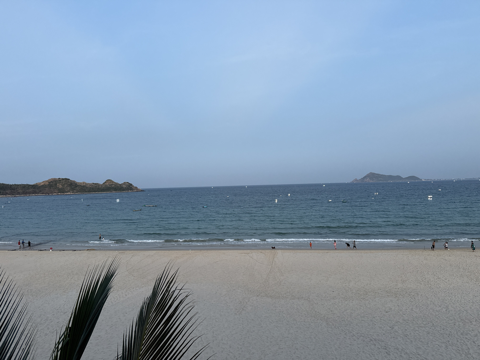 A wide, panoramic view of a tropical beach in Song Cau, Vietnam, unfolds under a pale blue sky. The foreground is dominated by a sandy beach, with a few fronds of a palm tree visible in the lower left corner, suggesting a natural, outdoor setting. The sand appears smooth and undisturbed in most areas, with faint tire tracks visible in the middle ground.
The middle ground features the calm, blue ocean, with gentle waves lapping at the shore. Several small boats are scattered across the water, some appearing to be fishing vessels. A few individuals are visible along the water's edge, engaged in various activities. On the left, a group of people stand near the water, while further to the right, a lone figure in an orange shirt walks along the shoreline. A dark-colored dog can be seen on the beach, possibly being walked by one of the beachgoers.
In the distance, two islands or peninsulas rise from the sea. The larger one on the left is covered in lush greenery, with its slopes leading down to the water. A smaller, more distant landmass is visible on the right. The overall atmosphere is serene and peaceful, characteristic of a coastal vacation spot in Song Cau.