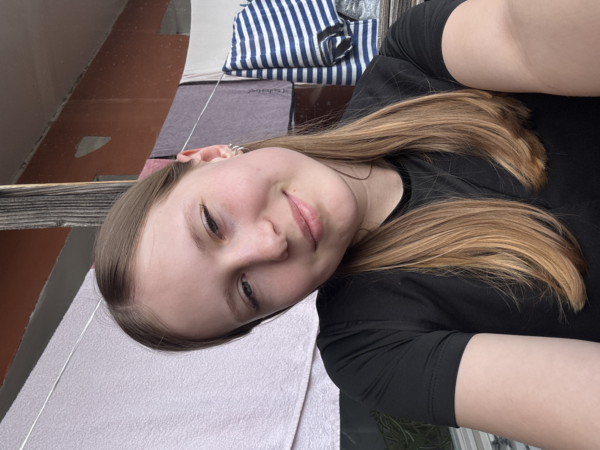 The image is an angled, medium close-up portrait of a young woman, likely a selfie, taken in Chaikovsky, Russia. The woman has light brown hair, fair skin, and light-colored eyes, and she is looking directly at the camera with a gentle smile. She is wearing a black short-sleeved top, and her left ear features multiple small silver hoop earrings.
Her hair frames her face and flows down across her shoulders. In the background, to the upper left, a reddish-brown wall or surface shows signs of wear with lighter patches. Below this, a dark, rustic-looking wooden or metal railing is visible. To the upper right, behind the woman's head, a blue and white striped fabric, possibly a towel or folded garment, is neatly stacked. Adjacent to this, a darker rectangular object features faint, illegible text that appears to be "CaraHeyPooT" or similar. A thin, silvery line, possibly a thread or wire, stretches diagonally across the image from the upper left, passing her ear and extending towards the foreground.
In the foreground and lower left, a light-colored, textured fabric, resembling a pink or white towel, is draped over a surface. The lighting suggests it is daytime, with soft ambient light. The setting appears to be a casual, possibly domestic outdoor or semi-outdoor space like a balcony.