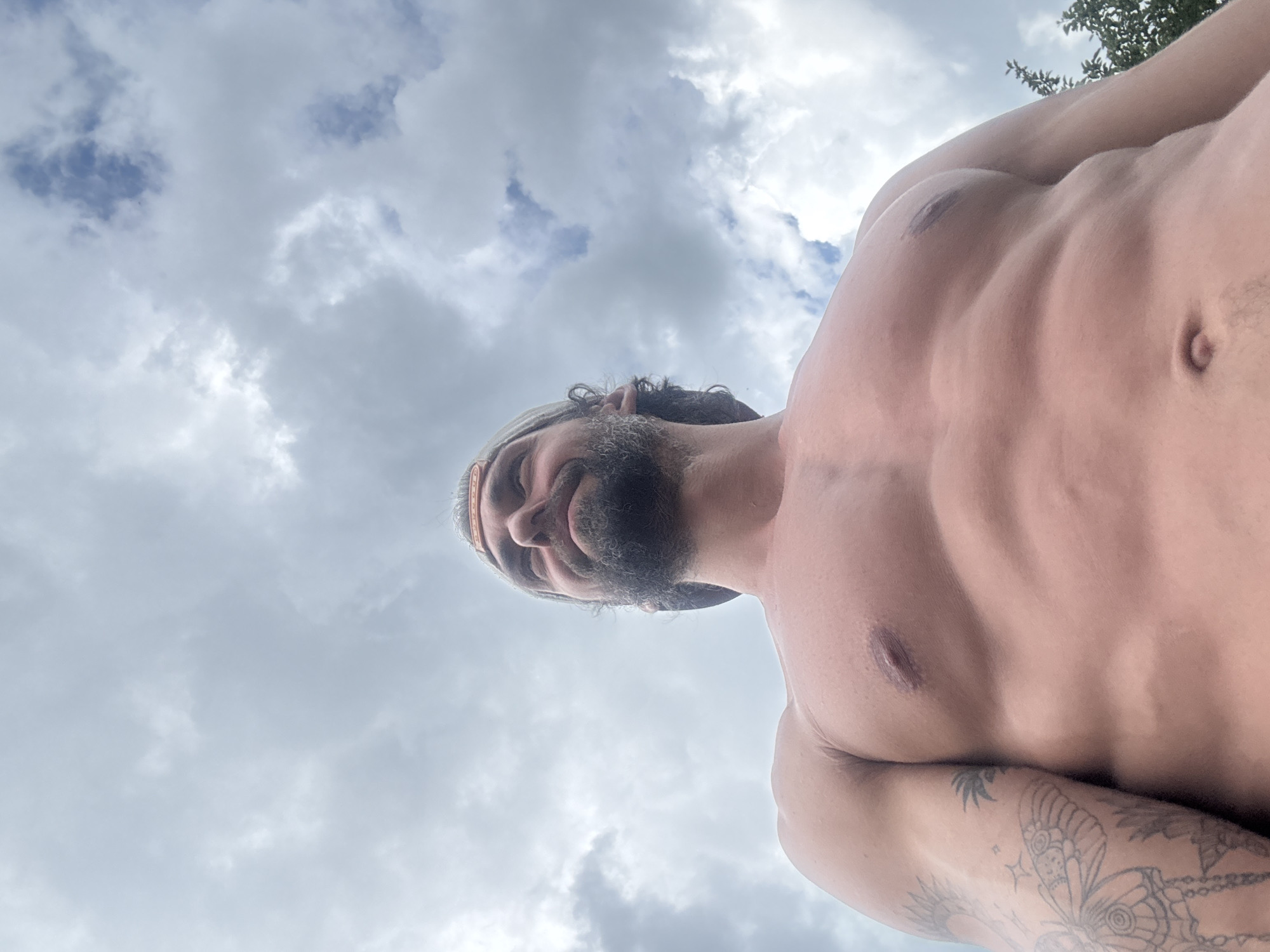 The media file shows a close-up, low-angle shot of a shirtless man against a cloudy sky. The man has a beard and is wearing a light-colored cap. His chest and abdomen are visible, displaying defined musculature and a tattoo on his arm. The sky is predominantly filled with gray and white clouds, suggesting overcast weather. The camera is positioned below the man, looking up at him, which creates a dramatic perspective. The lighting appears to be natural daylight, likely during the daytime. The image captures a moment of casualness, possibly outdoors given the sky and the man's attire. There is no visible text in the image.