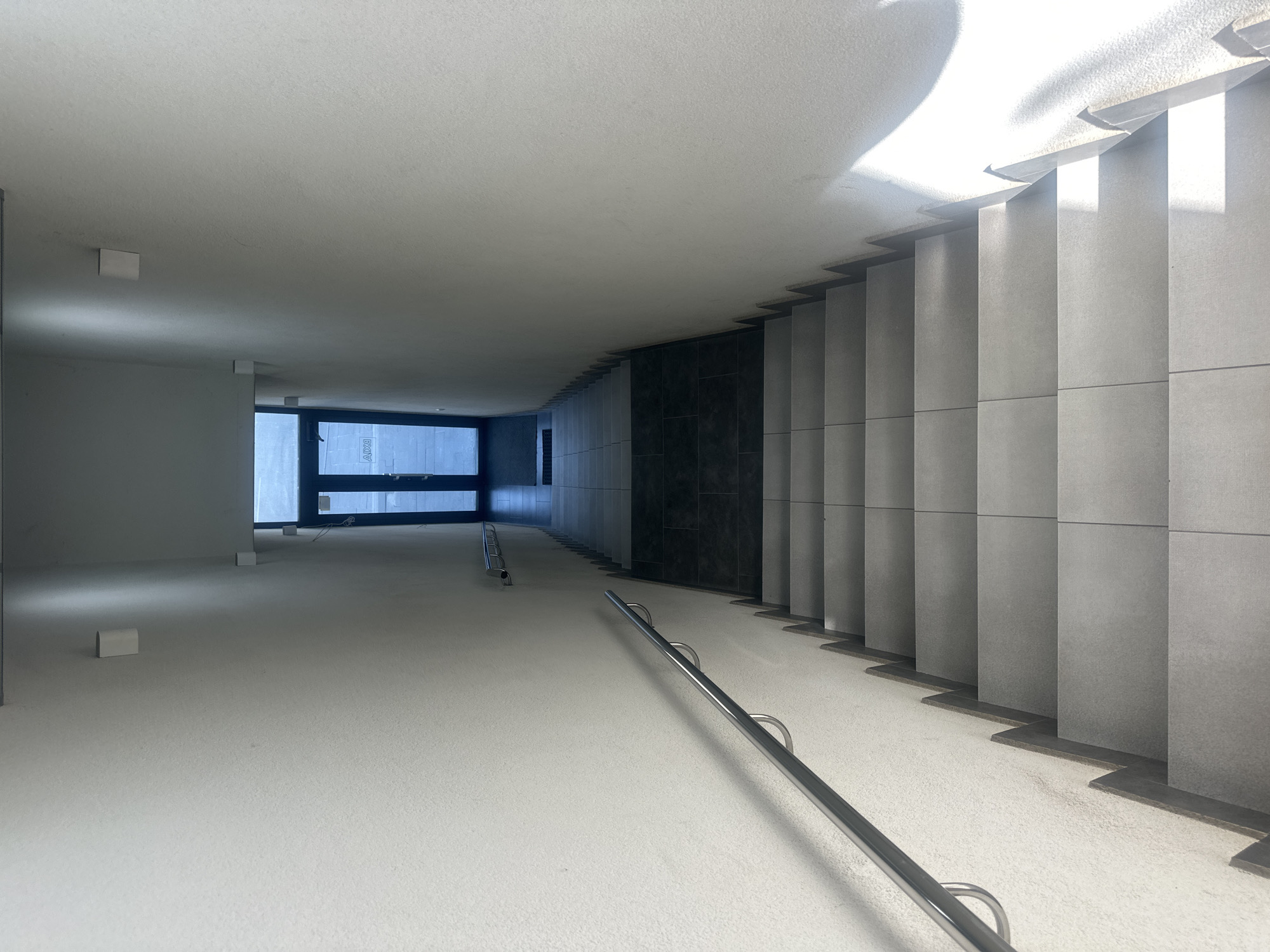 The image depicts an interior view of a modern, minimalist staircase and hallway in Ternopil, Ukraine. The scene is bathed in natural light streaming in from a large window or opening at the far end, suggesting it is daytime, likely with clear weather. The architecture is characterized by clean lines and neutral colors, primarily white and gray.
The primary subjects are the architectural elements themselves: a wide, gently sloping ramp or hallway leading to a staircase with concrete steps and gray tiled risers. A sleek, silver-colored metal handrail runs along the side of the ramp, curving upwards with the incline. The walls are painted white, and the ceiling is also white, with subtle shadows cast by the light.
In the distance, a large, dark-framed window or glass door reveals a glimpse of the exterior, with some light and faint shapes visible through it. There are a few small, white rectangular objects placed on the floor of the hallway, possibly construction materials or temporary markers.
The overall impression is one of quiet, uncluttered space, possibly a public building or a contemporary residential complex. There is no visible text in the image. The lighting creates a dramatic contrast between bright, sunlit areas and shadowed portions, highlighting the geometric shapes of the stairs and the space. The scene evokes a sense of calm and order.