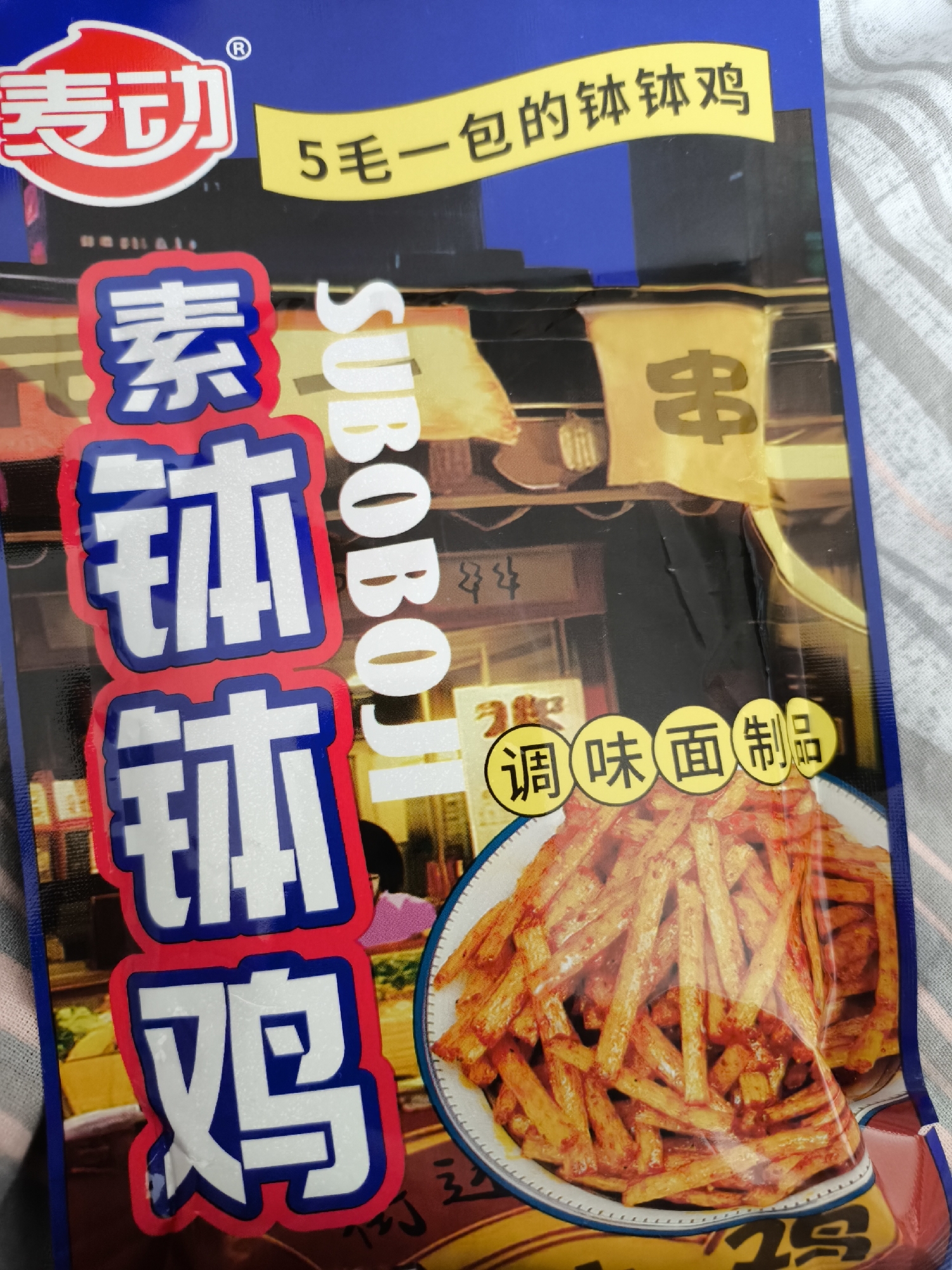 The image shows a package of a snack food product. The packaging is blue and features Chinese characters and the word "SUBOBOJ" in English. A photo of a plate of fried snacks is prominently displayed. A banner indicates the price is "5毛一包", suggesting it's inexpensive. The snack food is called "钵钵鸡". The environment around the image is a striped pattern.