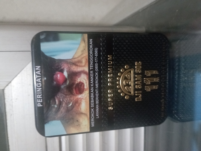 A vertically oriented, close-up photograph captures a "DJI SAM SOE" "SUPER PREMIUM" cigarette pack, likely resting on a flat surface in an indoor setting in Pubian, Indonesia. The pack is predominantly black with intricate gold-colored lettering and a textured appearance, featuring the brand name and "234" logo prominently.

The most striking detail is the graphic health warning covering a significant portion of the pack. This warning displays a disturbing image of a person suffering from severe throat cancer, showing exposed, raw tissue and a visible hole or stoma in the neck.

Accompanying the graphic are several lines of text in Indonesian. Vertically aligned on the left side of the warning is "PERINGATAN" (Warning). Below the graphic, the text reads: "MEROKOK SEBABKAN KANKER TENGGOROKAN" (Smoking causes throat cancer), followed by "LAYANAN BERHENTI MEROKOK (0800-177-6565)" (Quit Smoking Service (0800-177-6565)). The top right also indicates "SUPER PREMIUM".

The scene is static, focusing solely on the cigarette pack and its stark message against the backdrop of a light-colored, possibly reflective surface. No specific activity is depicted, and the time of day is not discernible.