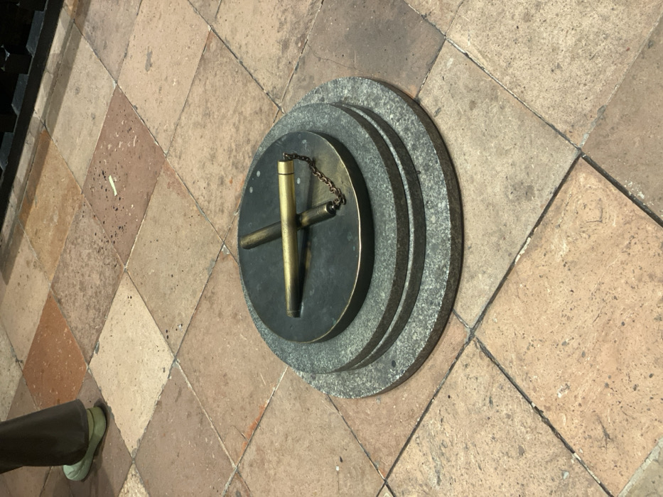 The image presents a high-angle view of a metallic, possibly brass or bronze, object resting on a tiered circular base, set into a tiled floor. The central object consists of a main vertical cylindrical piece with a short chain attached to its top. Two shorter, horizontal cylindrical bars extend perpendicularly from the base of the main cylinder, forming a cross shape. This assembly is placed on a dark, circular disk, which is then supported by two wider, concentric rings of light gray, speckled material resembling granite.
The entire structure is situated on a floor composed of square tiles laid out in a diamond pattern. These tiles vary in color from light brown to reddish-brown, showing some wear and texture. In the bottom left corner, the lower part of a person's leg, dressed in dark trousers and a light green shoe, is visible, indicating human presence.
The object appears to be a stationary fixture, possibly a historical or scientific instrument like a gnomon for a sundial, or a decorative marker. No specific activity is taking place beyond its static display. The lighting is even, suggesting an indoor setting or a well-lit covered area. Given the location context of Guang Zhou Shi, China, this could be part of a museum exhibit, a historical landmark, or a public space. There is no visible text in the image.